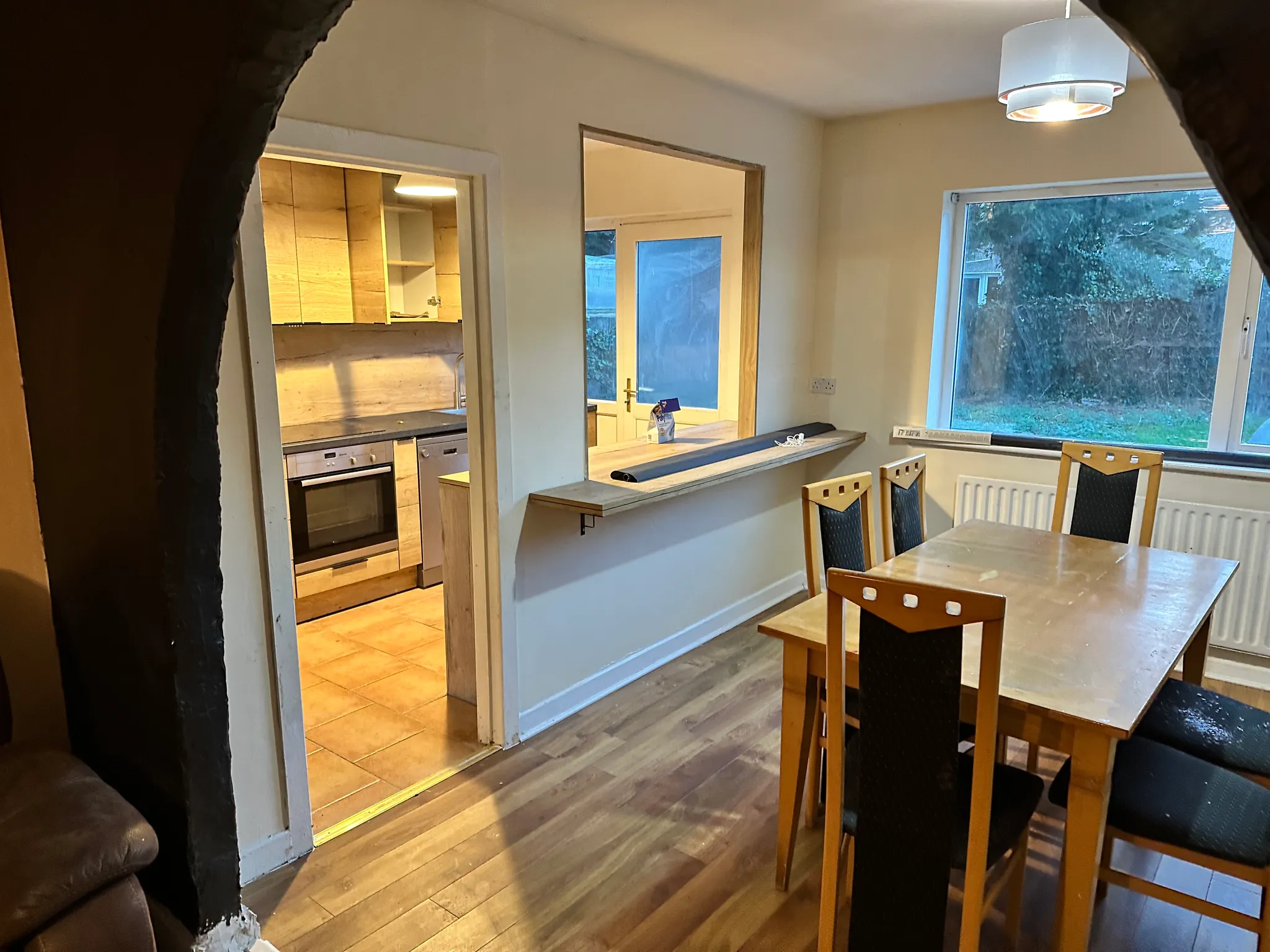 Dining room with a wooden table and four chairs, large window on the right, pendant light, and a passthrough opening showing an adjacent kitchen.