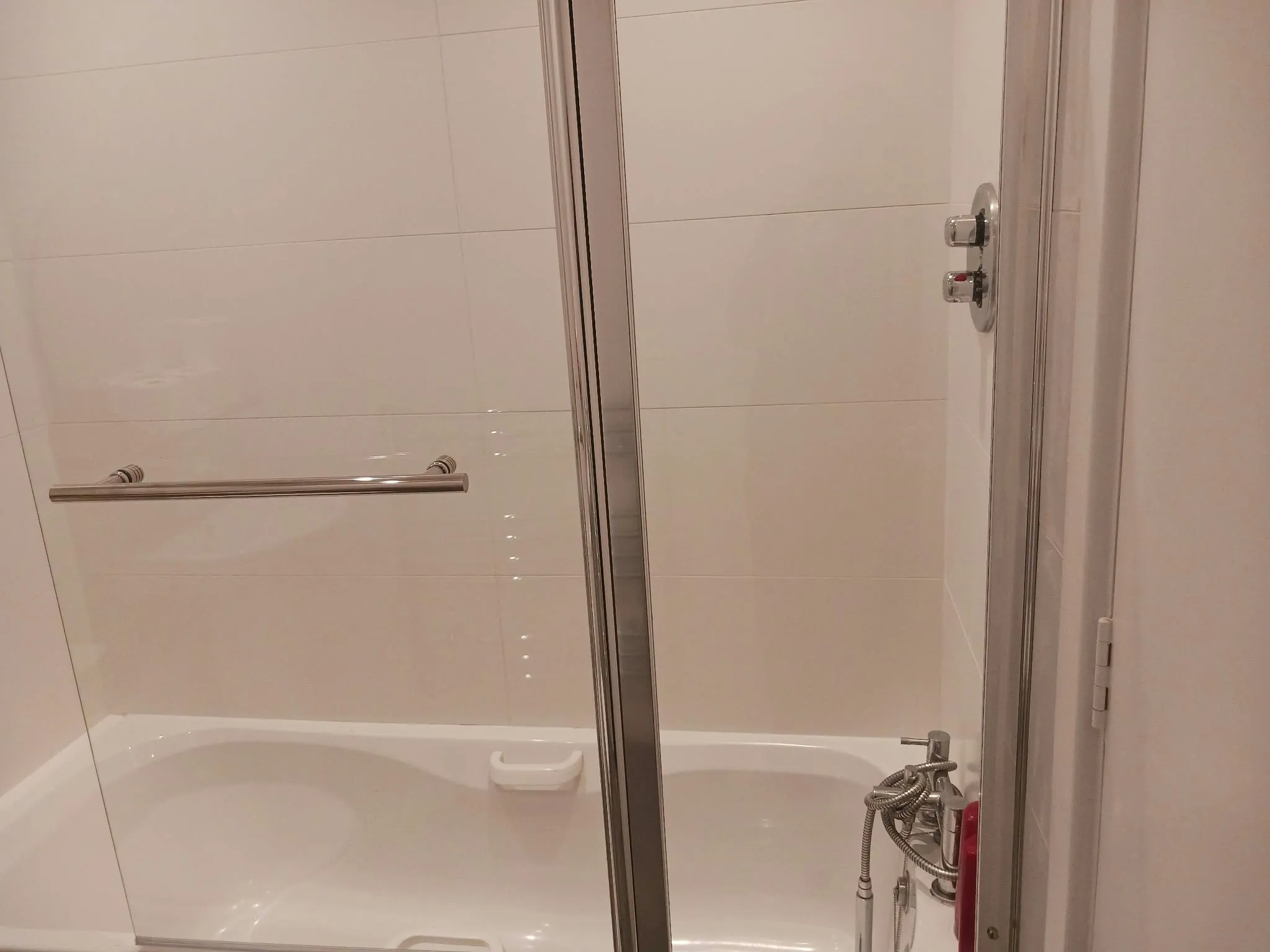Bathroom with a bathtub and glass shower door, chrome towel bar and handheld shower fixtures on tiled wall.