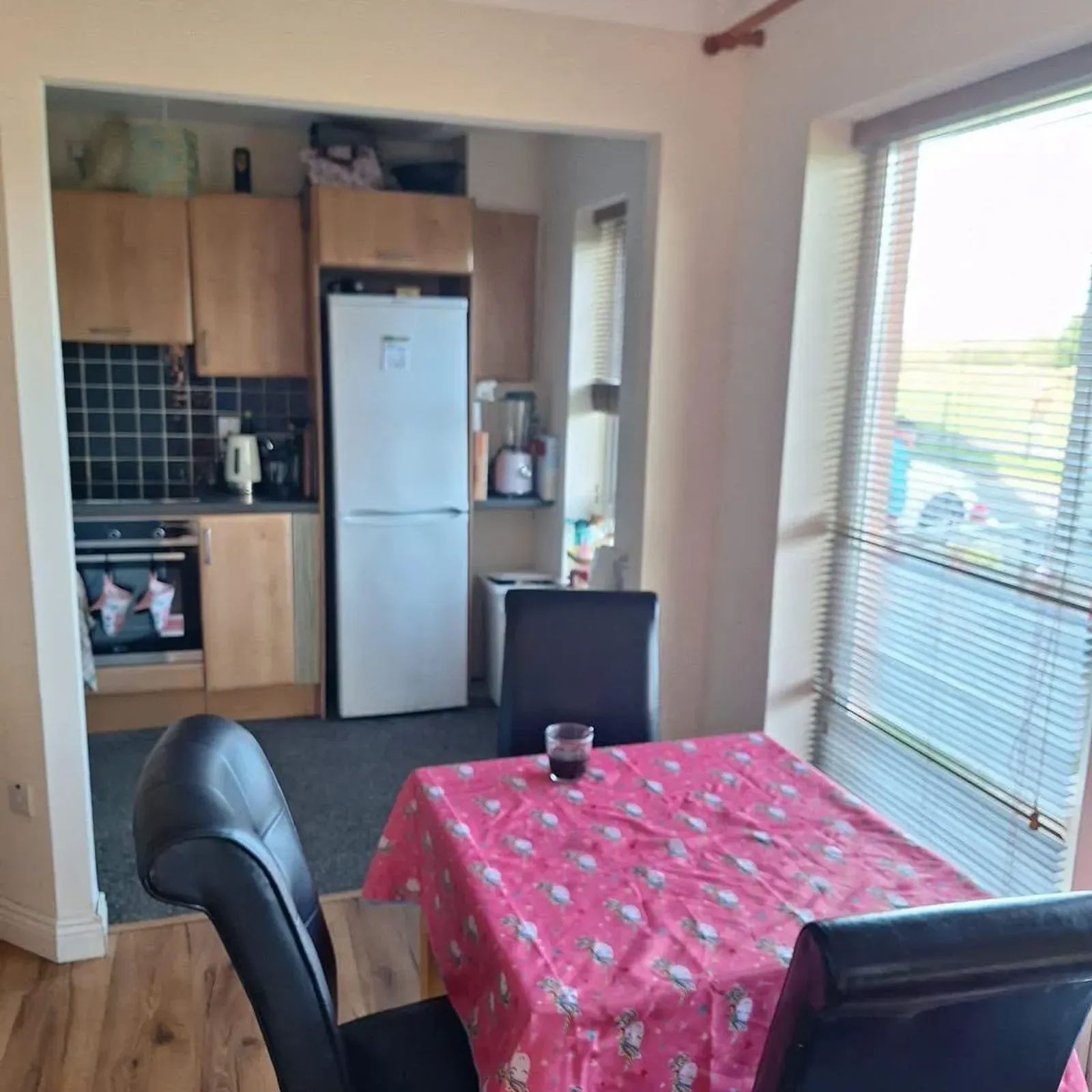 Dining area with a pink tablecloth on a rectangular table, several black dining chairs, a large window with blinds, and an open view into a small kitchen with a refrigerator and wooden cabinets.