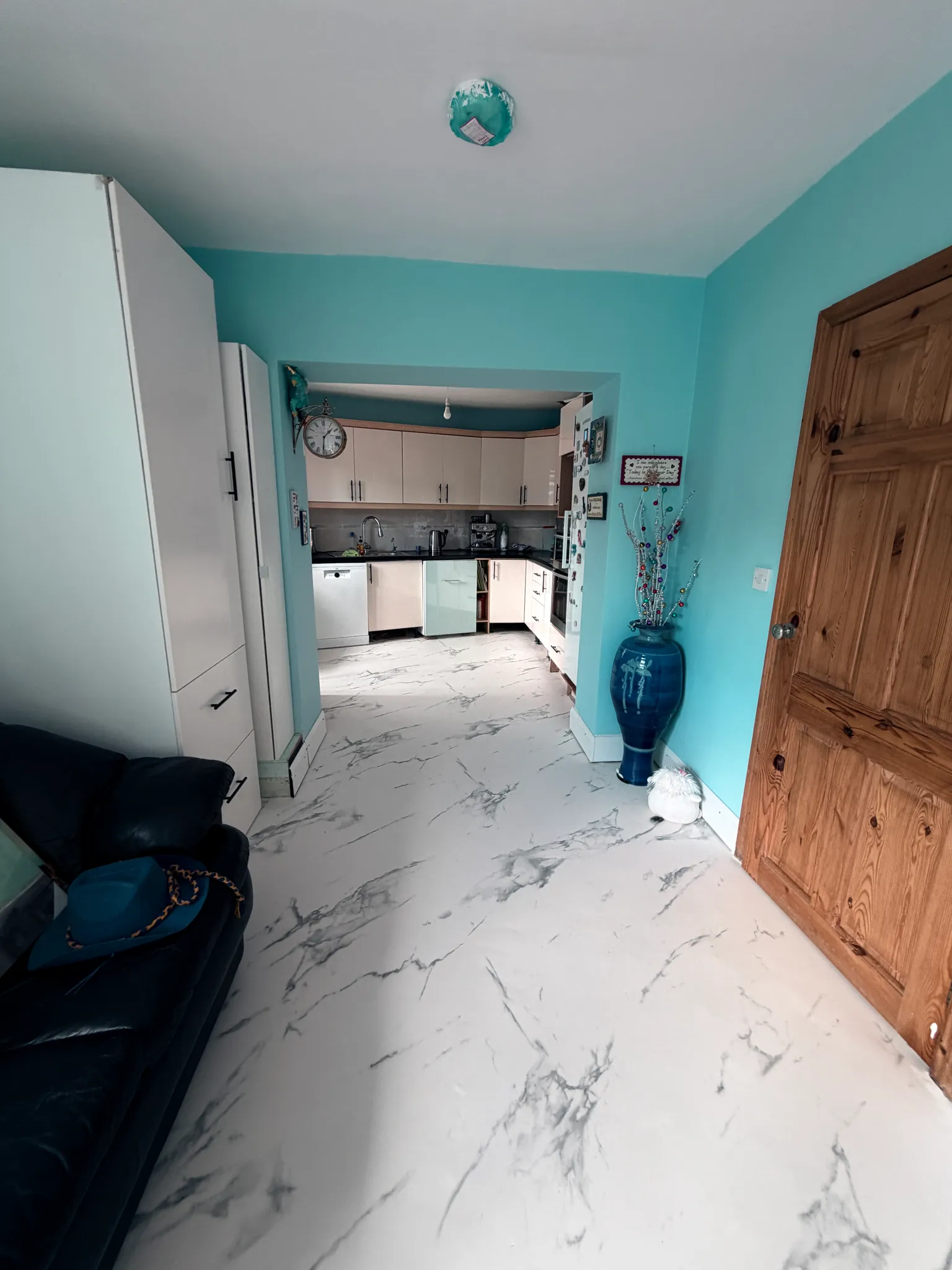 Open-plan view from a living area into a kitchen with turquoise walls, wooden door on the right, white cabinets and a dishwasher visible through a wide doorway.