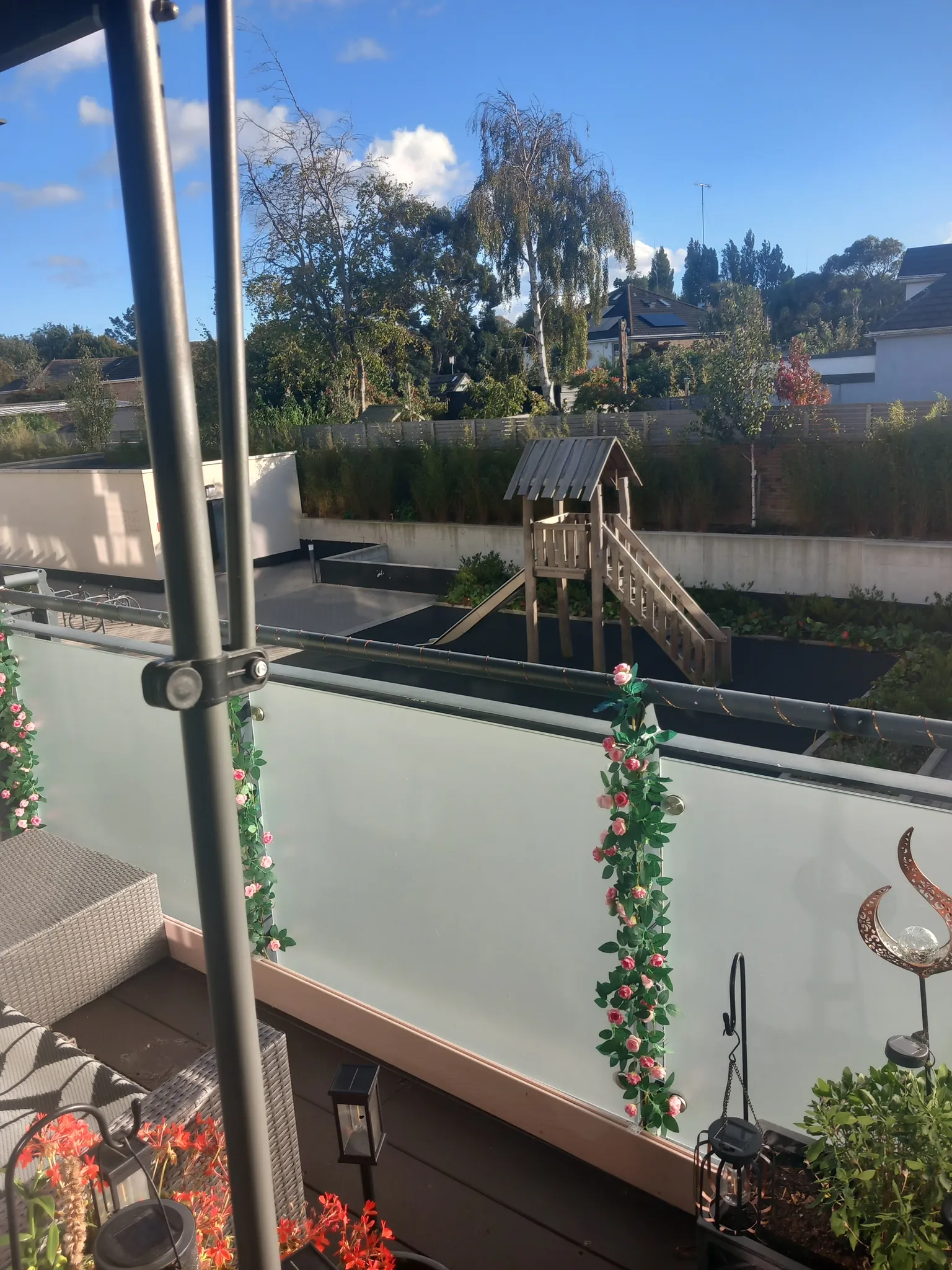 Balcony view with frosted glass railing decorated with artificial flower garlands, potted plants and lanterns; a backyard with a wooden play structure, trees and houses under a blue sky.