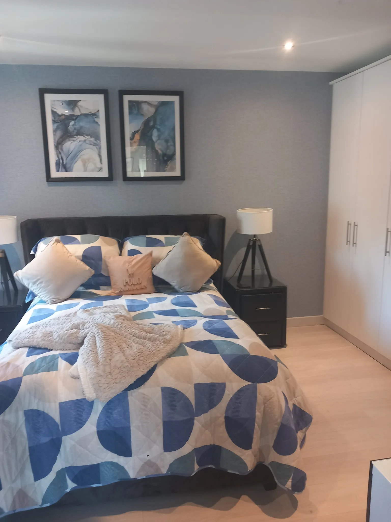 Bedroom with a double bed and patterned bedspread, two framed prints above the headboard, a nightstand with a tripod lamp, and a white wardrobe on the right.