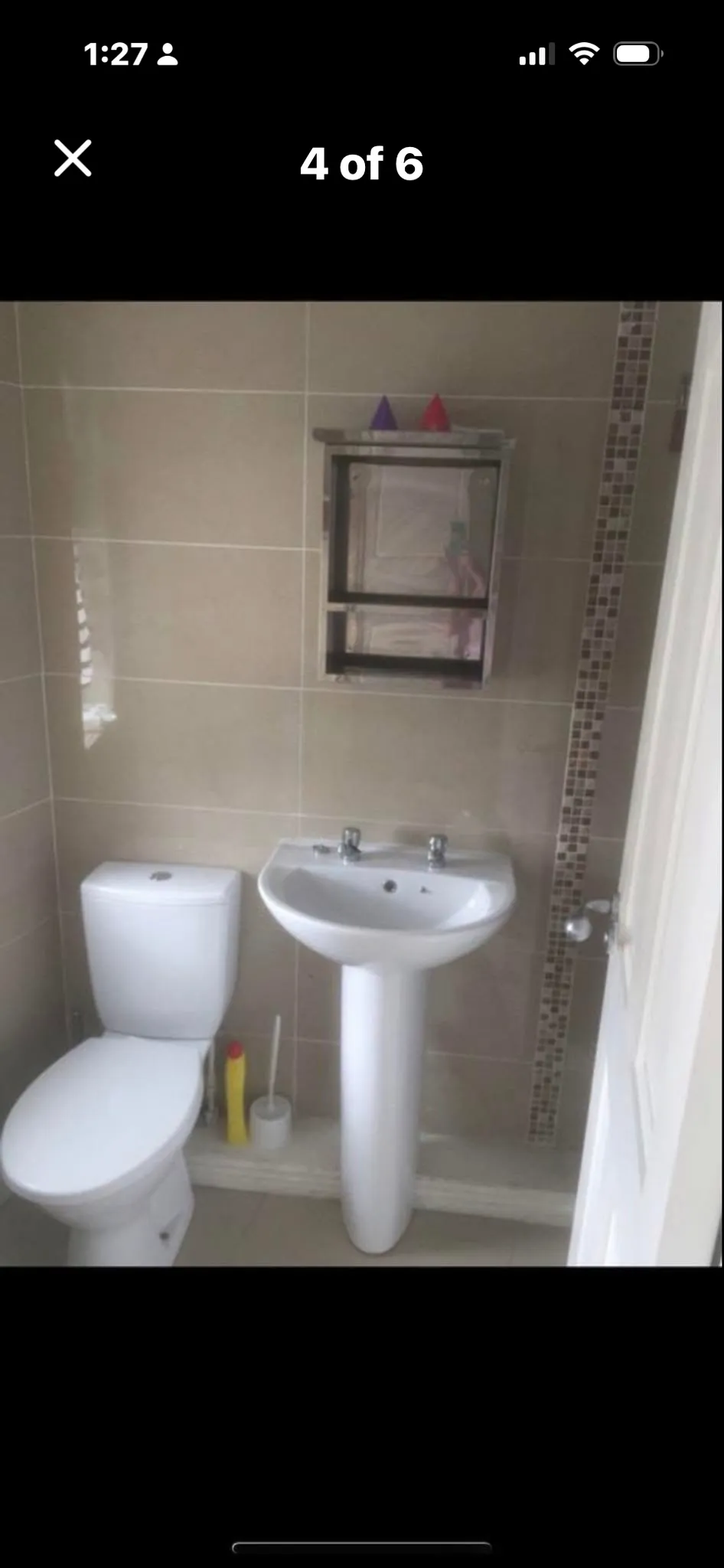 Small bathroom with a toilet, pedestal sink with two taps, and a mirrored medicine cabinet mounted on tiled wall with a vertical mosaic tile strip.
