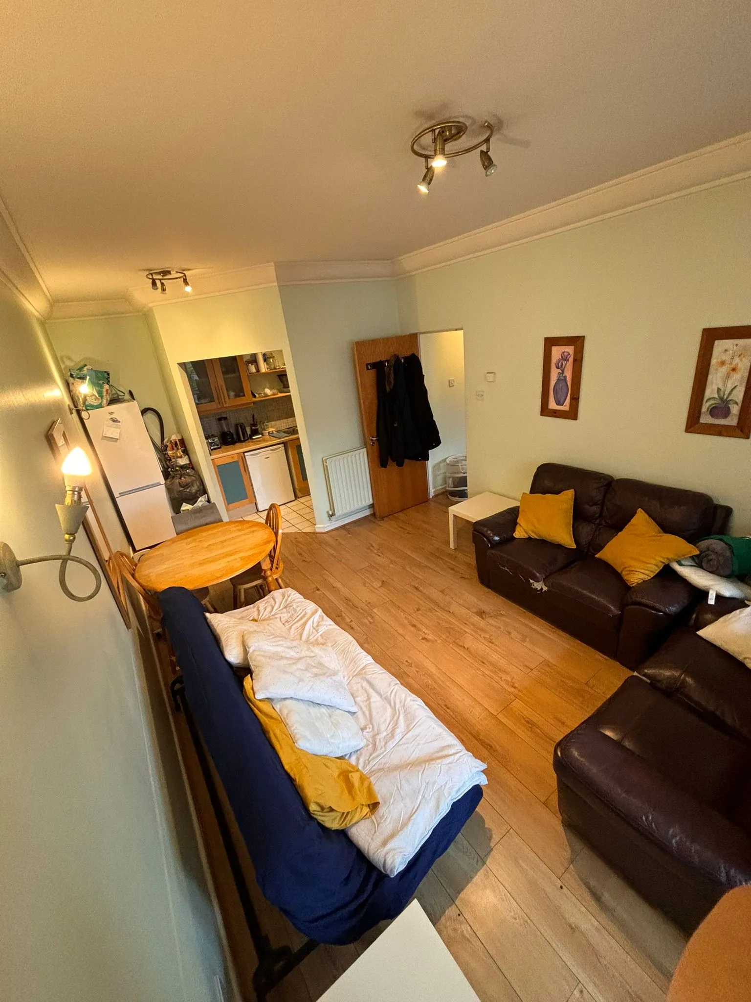 Living room with a sofa and yellow cushions, a small white coffee table, a round wooden dining table, a futon or daybed along one wall, and a kitchenette visible through an open doorway.