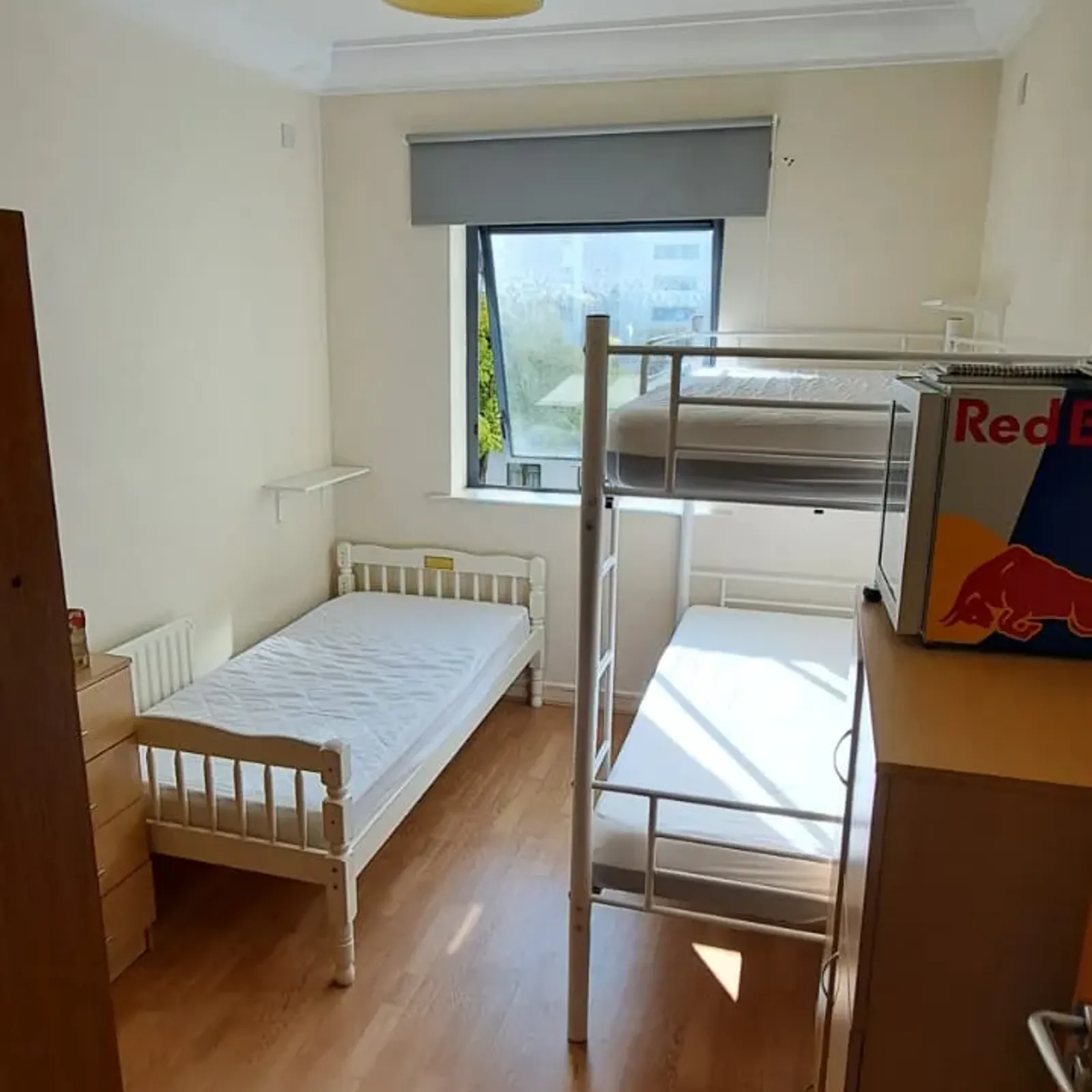Bedroom with a single bed and a bunk bed, window with a roller blind, wooden floor, dresser and ceiling light.
