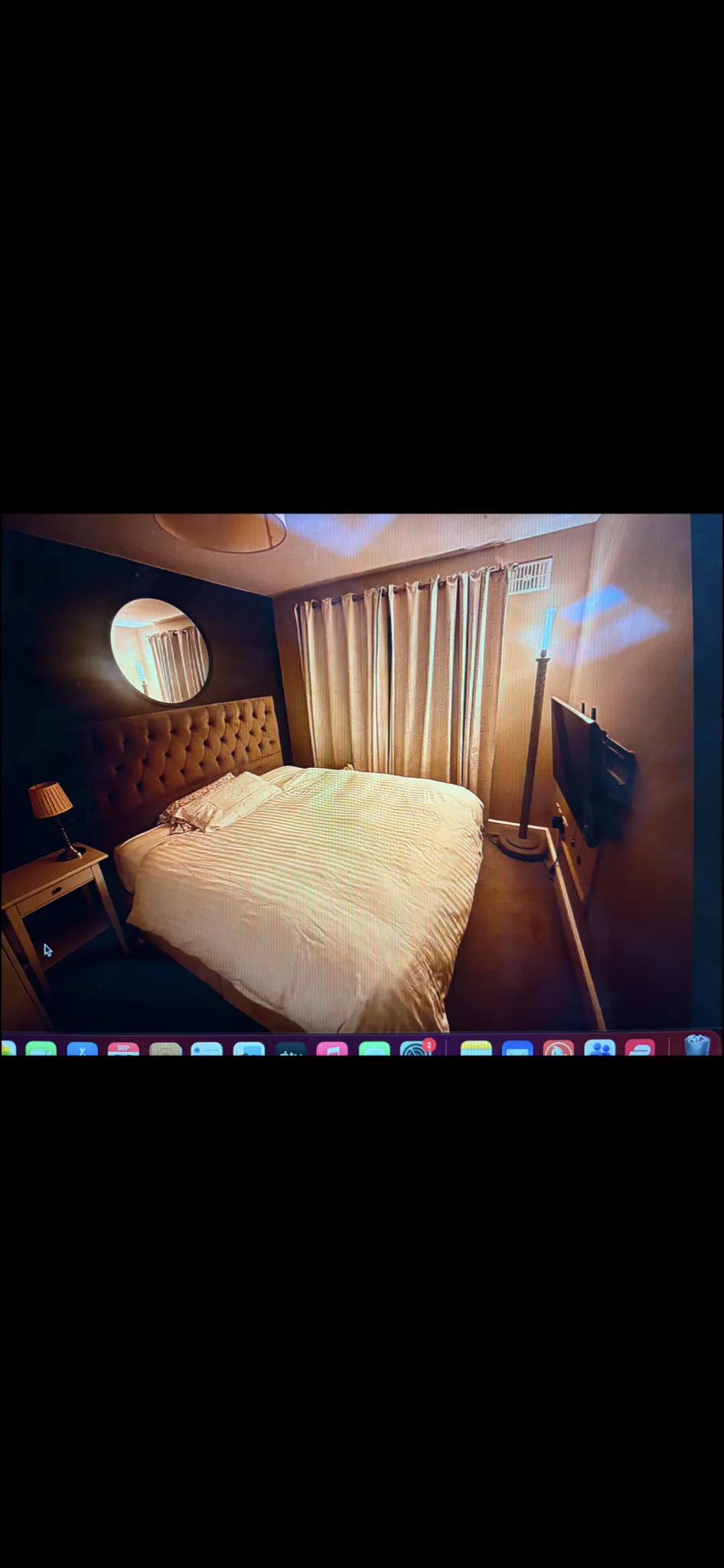 Bedroom with a double bed, tufted headboard, striped bedding, round mirror, bedside lamp, floor-to-ceiling curtains, and wall-mounted TV