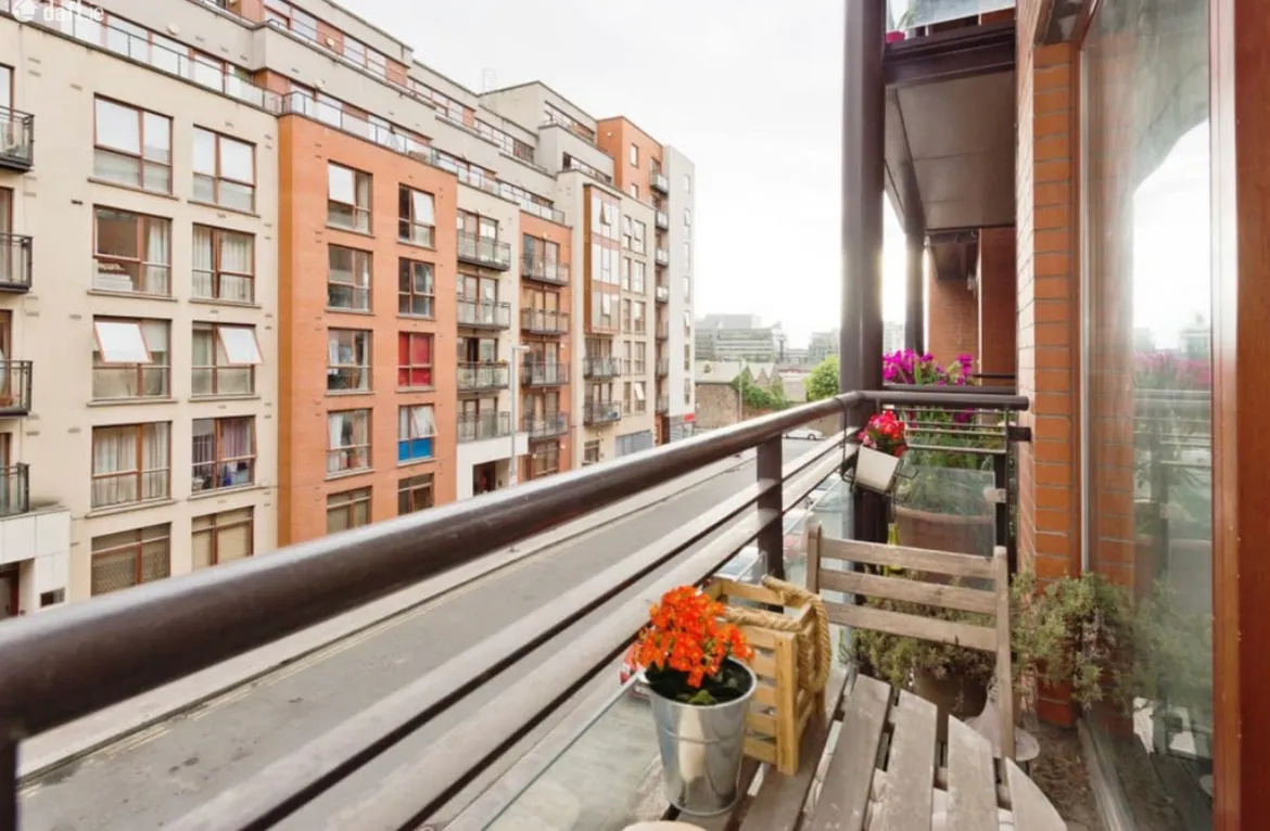 Balcony with a metal railing, small wooden table and chairs, potted flowers and a view of neighboring apartment buildings across the street.
