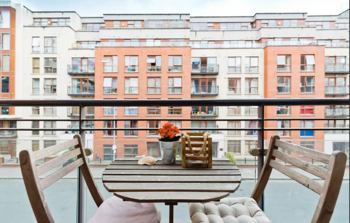 Small balcony with a round wooden table, two wooden chairs with cushions, a potted flowering plant and a city apartment building visible across the street