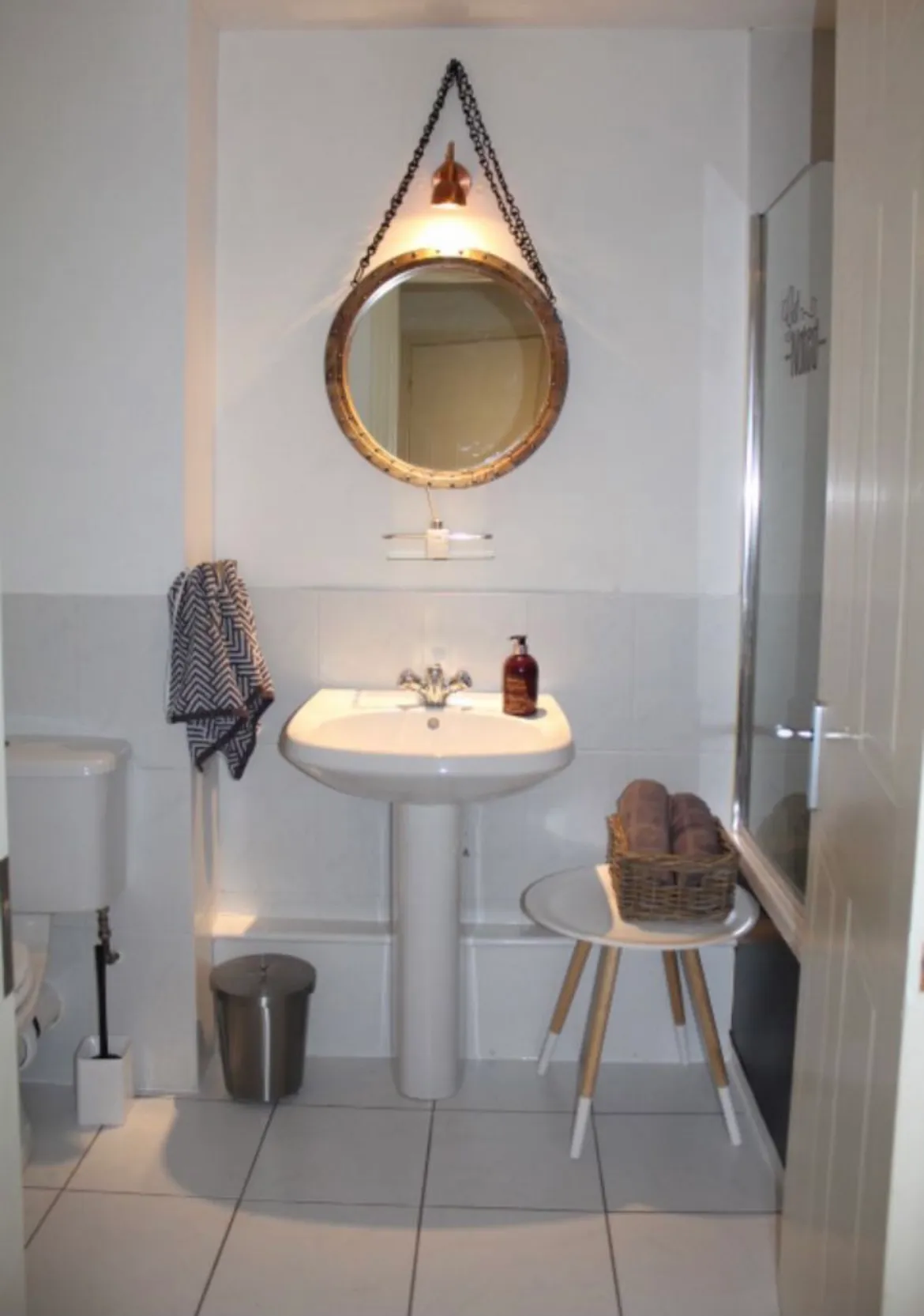Bathroom with a pedestal sink, round hanging mirror, towel on a hook, soap dispenser, and a small stool holding a basket of towels next to a shower door.