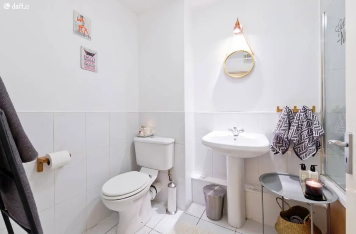 Bathroom with a toilet, pedestal sink, round wall mirror, towel hooks, and a small side table with toiletries and a candle.