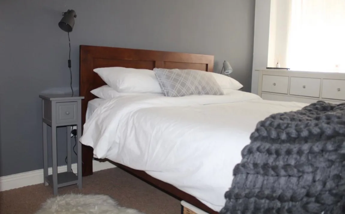 Bedroom with a wooden headboard bed made with white bedding, a gray throw blanket, bedside table with lamp, and a dresser by a bright window.
