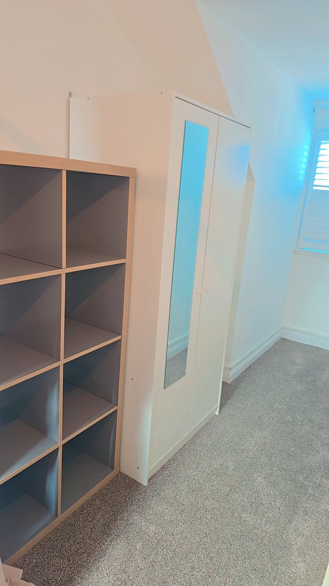 Bedroom or workspace with wooden shelving unit on left wall, white wardrobe with mirror doors on right, gray carpet flooring, and bright natural light from window