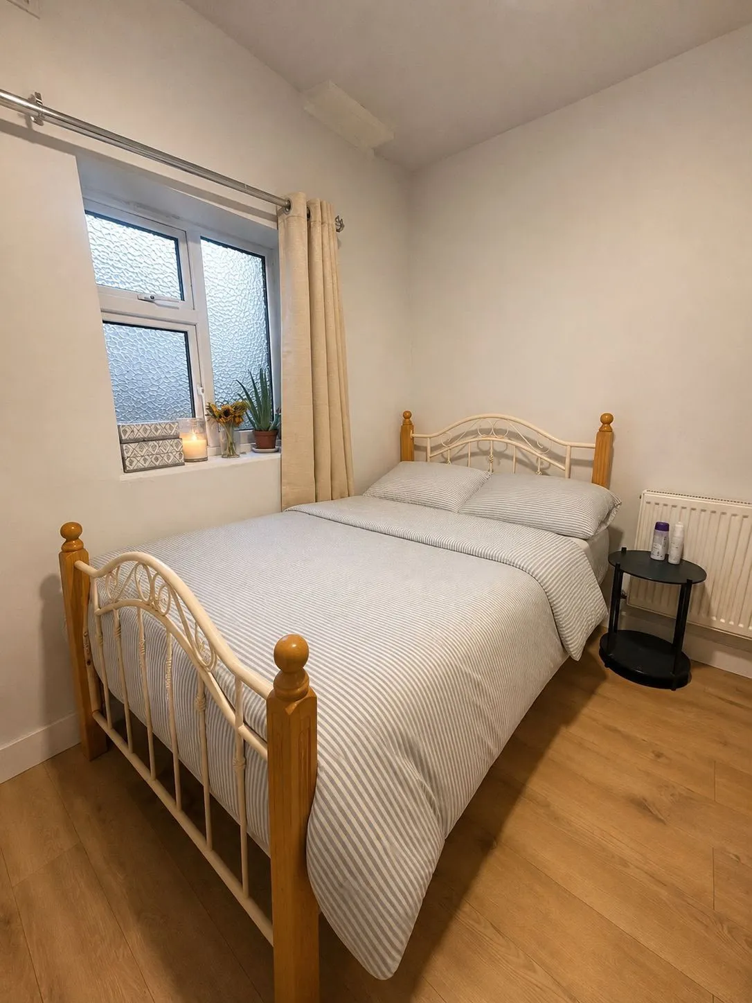 Bedroom with a double bed against a white wall, a window with frosted glass and curtains, a small round bedside table, and a radiator.
