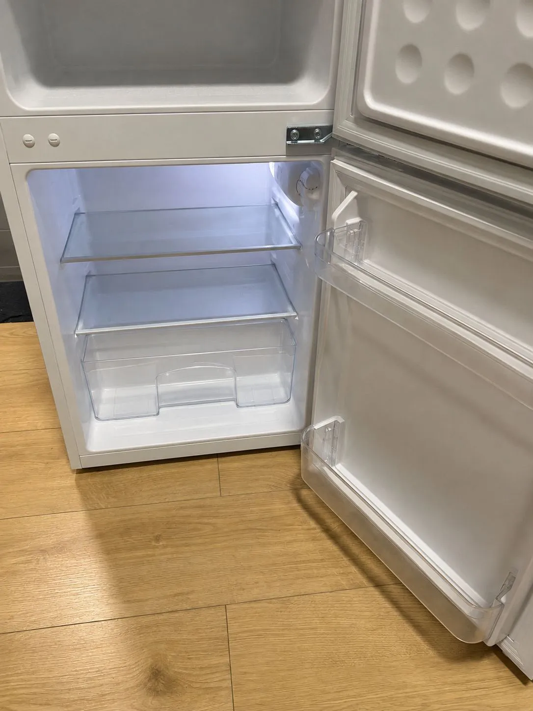 Open refrigerator with empty interior shelves, a clear crisper drawer and door shelves visible, standing on a wood floor.