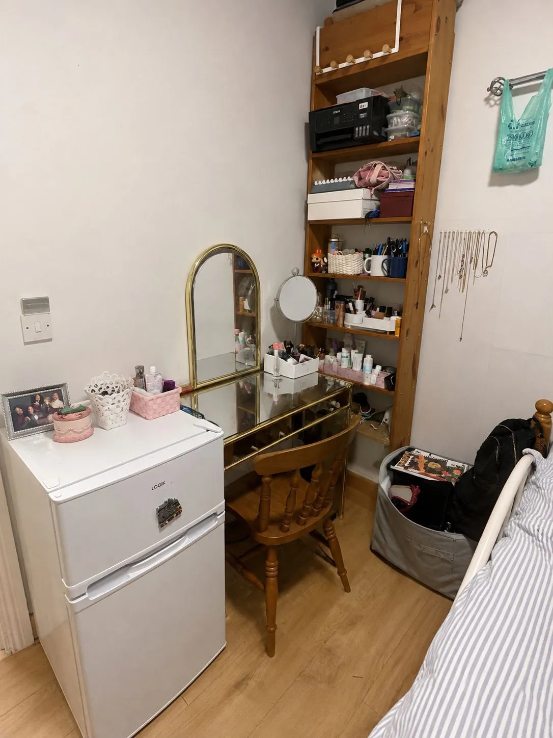 Bedroom corner with a dressing table and mirror, wooden chair, tall open shelving with toiletries and boxes, a small white refrigerator, and the edge of a bed.
