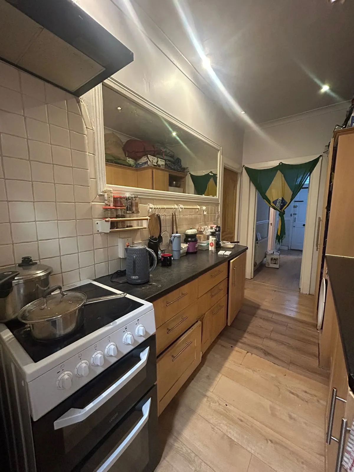Narrow kitchen with a freestanding stove, pots on the hob, a countertop with kettle and small appliances, wooden lower cabinets, tiled backsplash and a large wall mirror, view toward a hallway with fabric hung in the doo