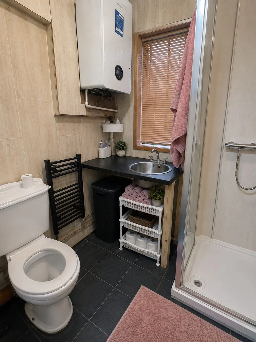 Bathroom with a toilet, sink set into a black countertop, corner shower, wall-mounted boiler above the counter, window with blinds, shelving holding rolled towels and toilet paper, and a pink bathmat.