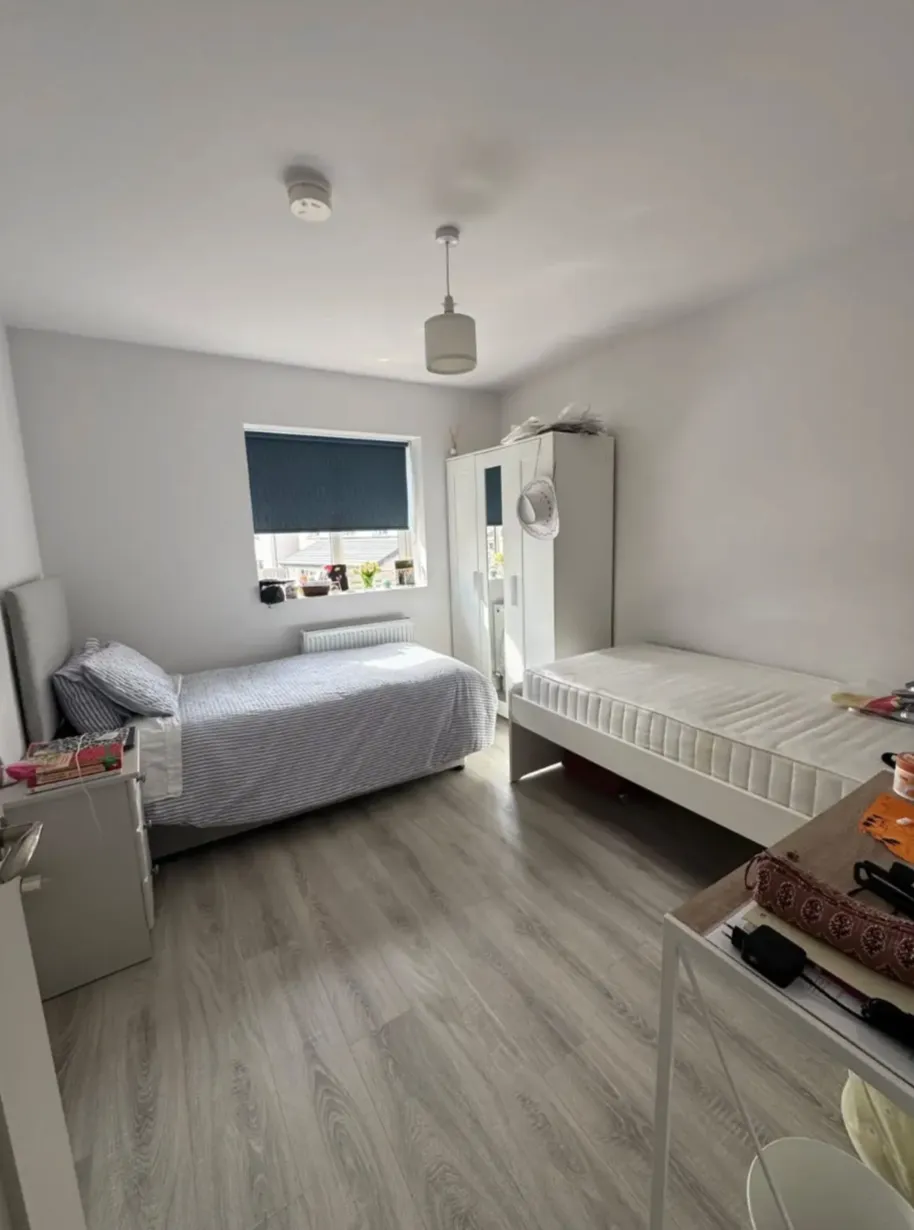 Bedroom with two single beds, a wardrobe with mirror, a window with a dark blind, a small bedside table and a desk, with light wood laminate flooring.