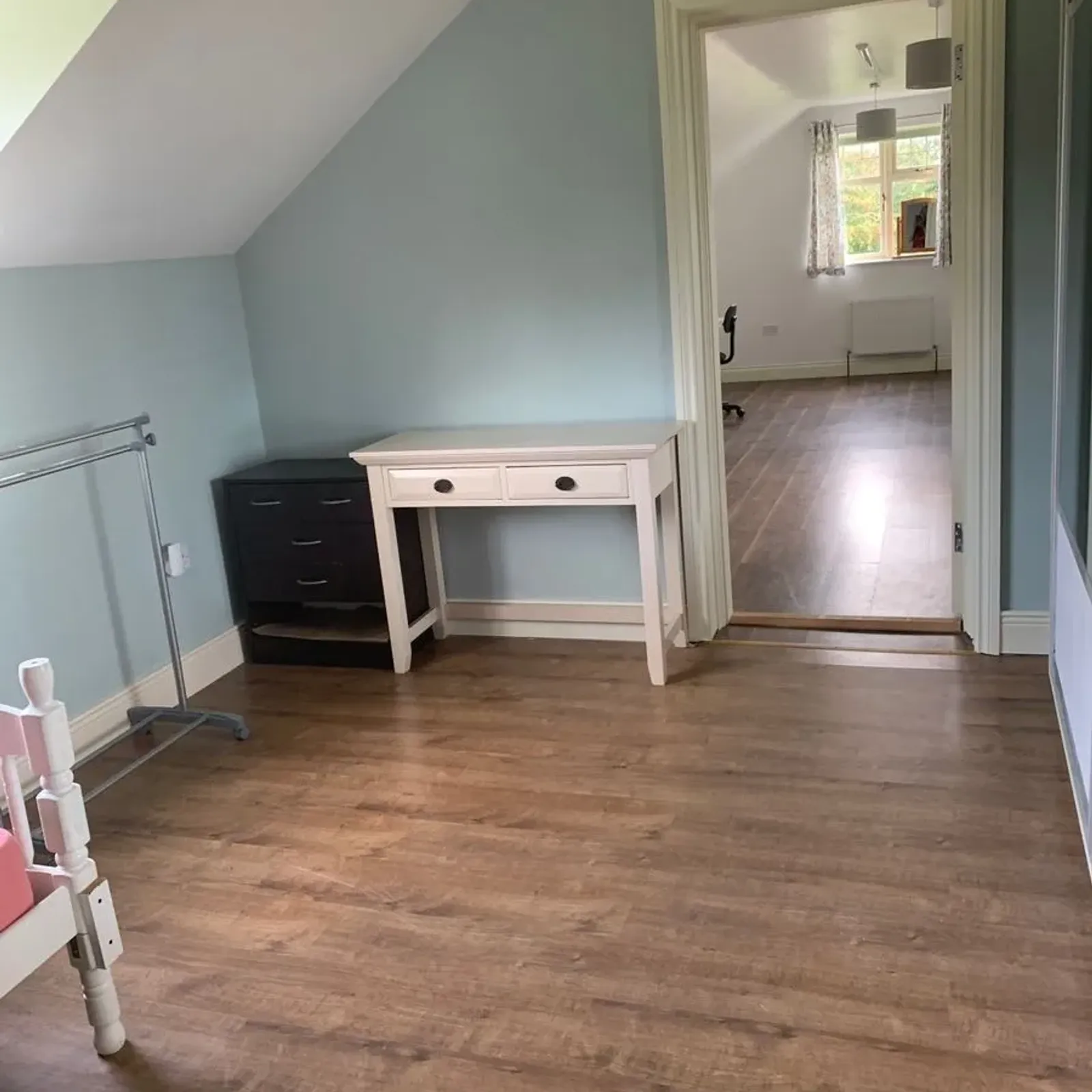 Bedroom with wooden floor, small desk, chest of drawers, clothes rail and doorway leading to another room with a window.