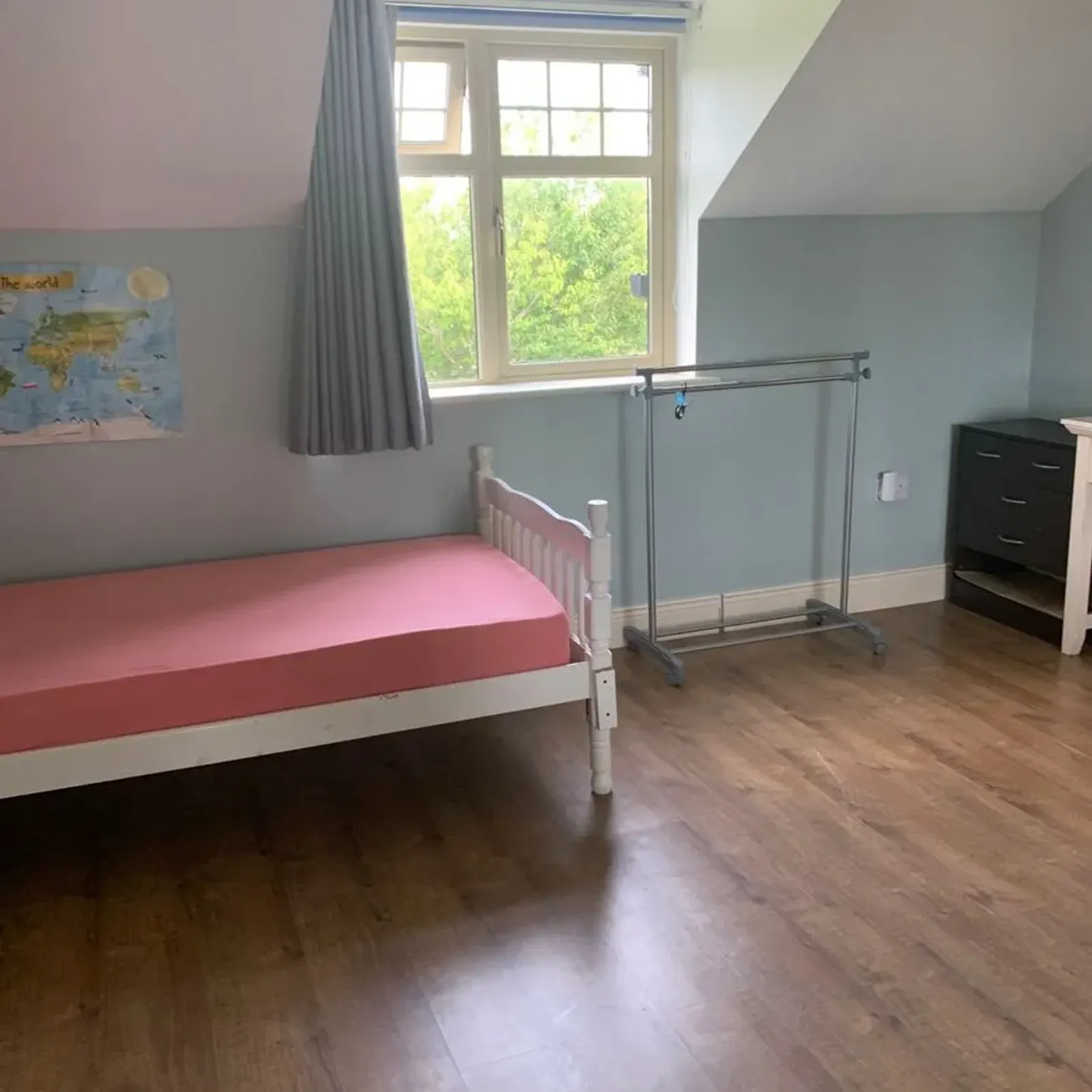 Bedroom with a single bed with pink mattress, window with curtain, freestanding clothes rail and wooden floor