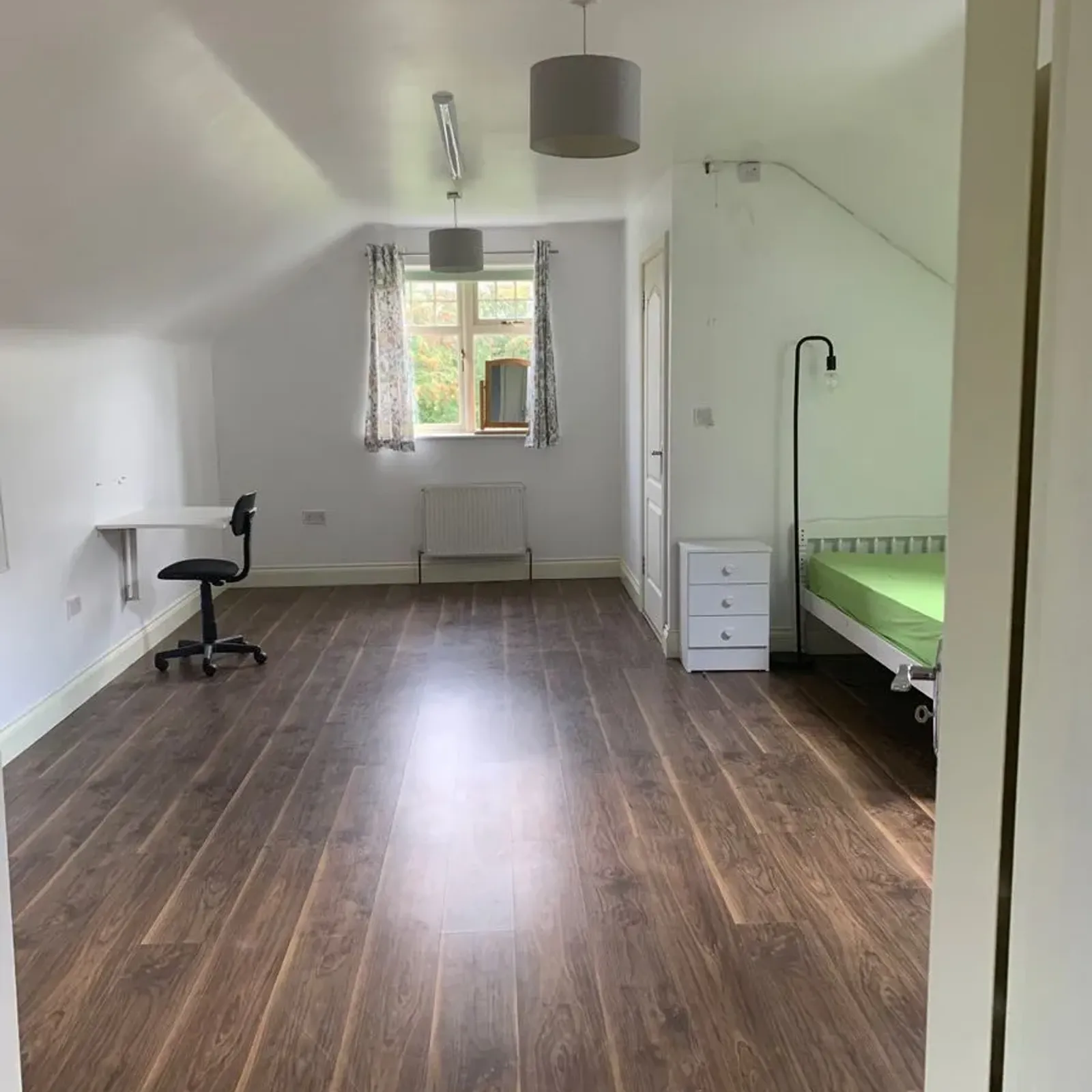 Bedroom with wood-look floor, single bed with green mattress and bedside drawer on the right, a floor lamp, a window with curtains and a small wall-mounted desk with an office chair on the left.