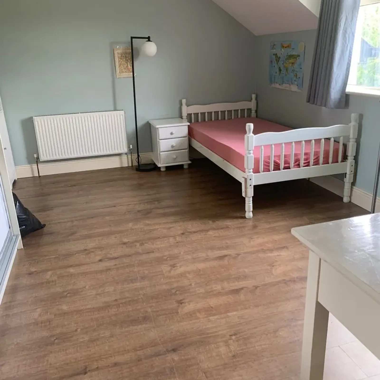 Bedroom with a single bed, pink mattress, white nightstand and floor lamp near a window with grey curtain, radiator on pale blue wall and wood-look floor.