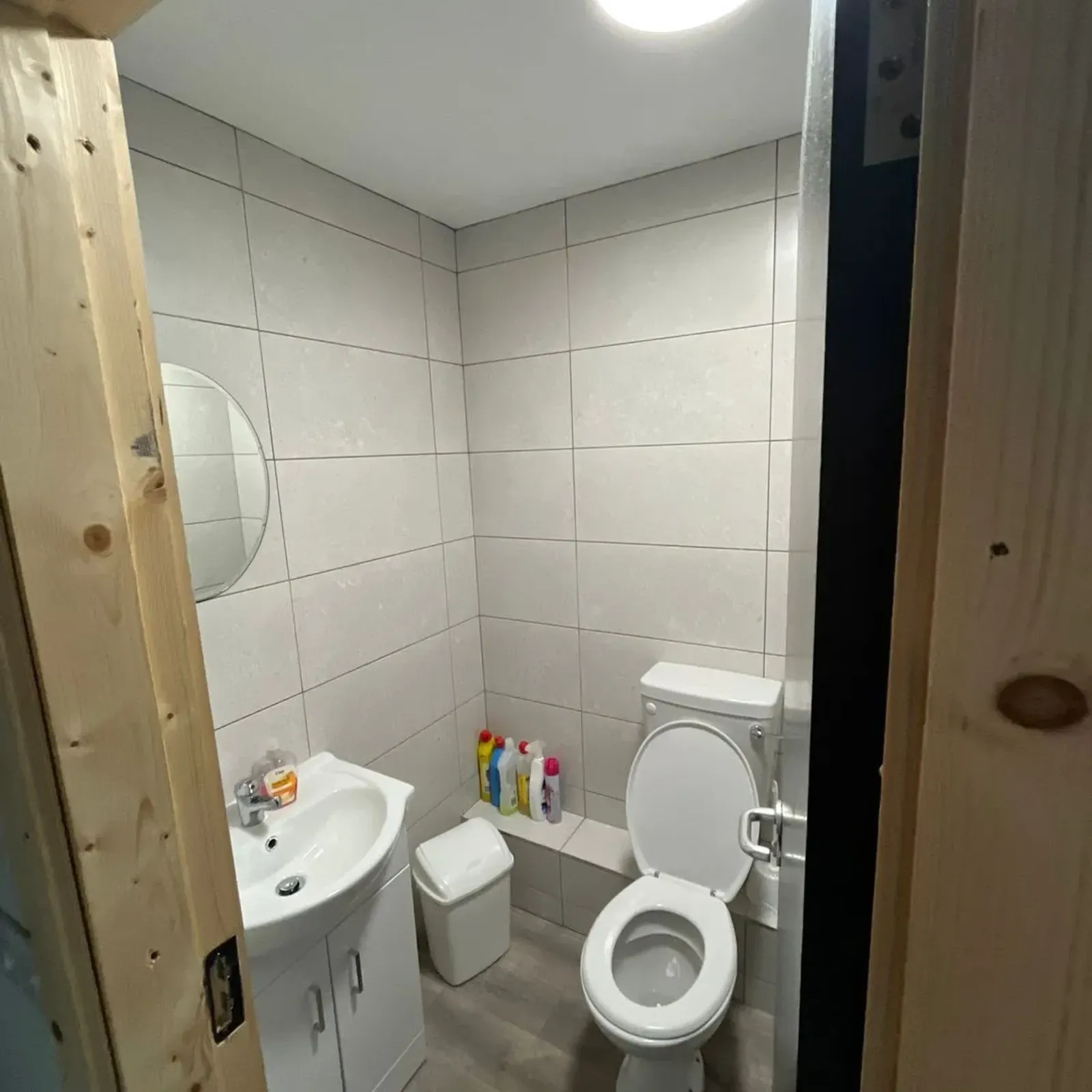Small bathroom with a toilet (seat up), sink with cabinet, round wall mirror, tiled walls, ceiling light and cleaning products on a ledge.
