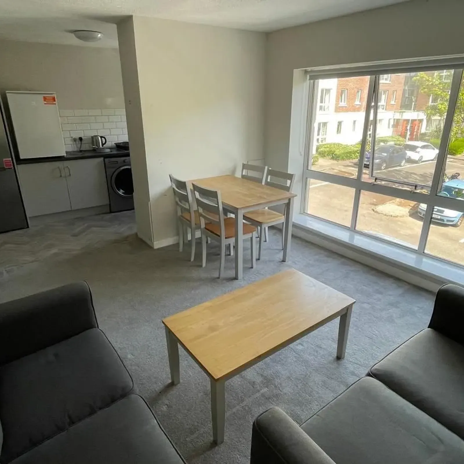 Open-plan living room and kitchenette with two sofas, a small wooden coffee table, a dining table with four chairs, large front window overlooking a parking area, and visible fridge and washer in the kitchen area.