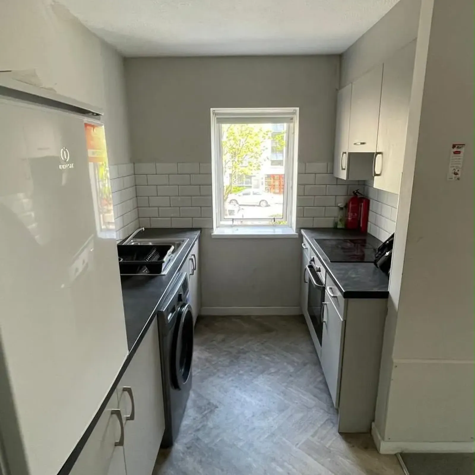 Narrow kitchen with countertops on both sides, sink and washing machine on the left, oven and cabinets on the right, small window at the far end and ceiling light