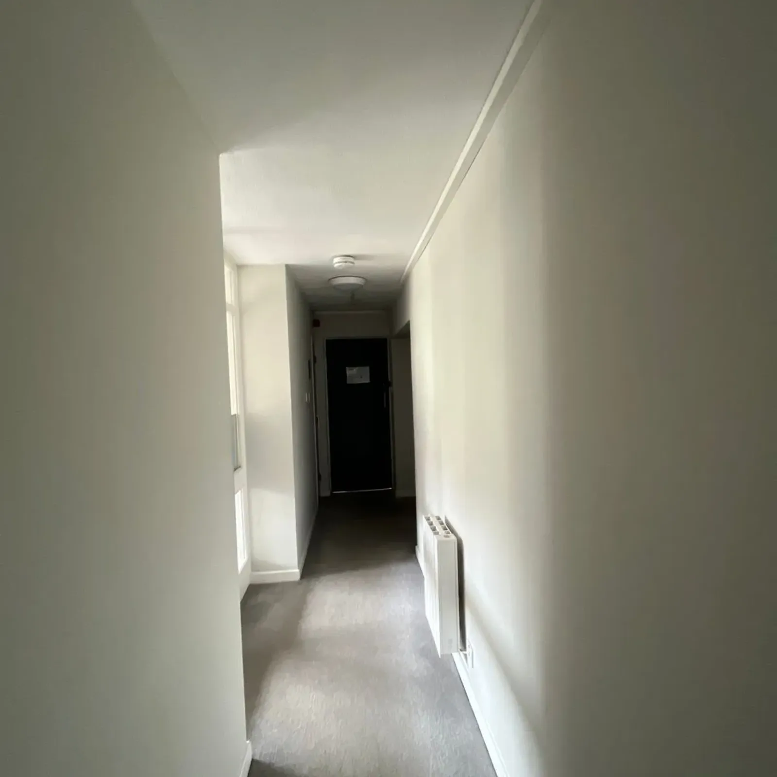 Narrow hallway with carpeted floor, a radiator on the right wall and a door at the far end