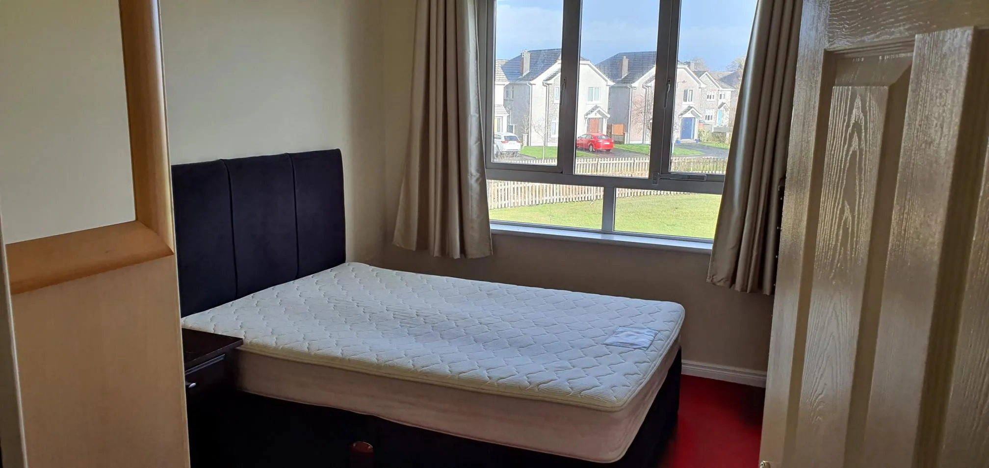 Bedroom with a single bed with black headboard and white mattress, large window with view of residential street and neighboring houses, beige curtains, wooden wardrobe on right side, red carpet flooring.