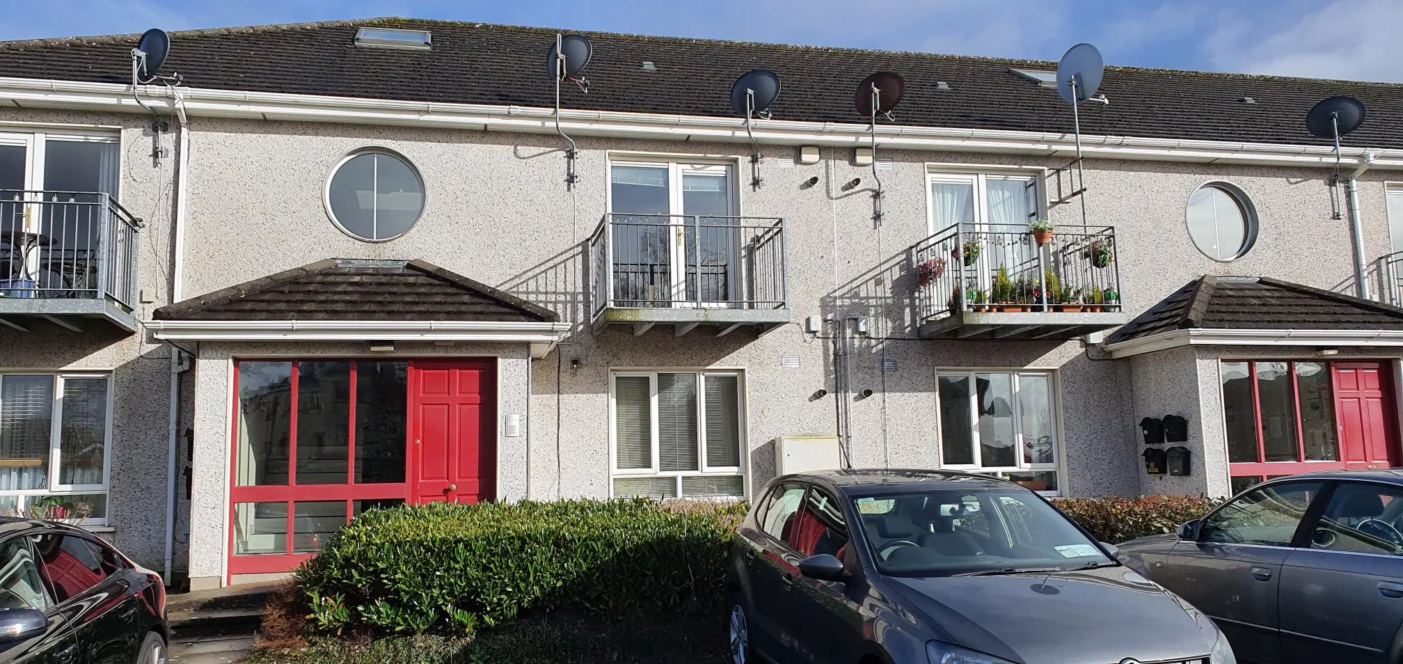 Exterior of a multi-unit residential building with cream-colored render, dark roof tiles, multiple balconies with metal railings, circular windows, red entrance doors, satellite dishes, and parked cars in front.