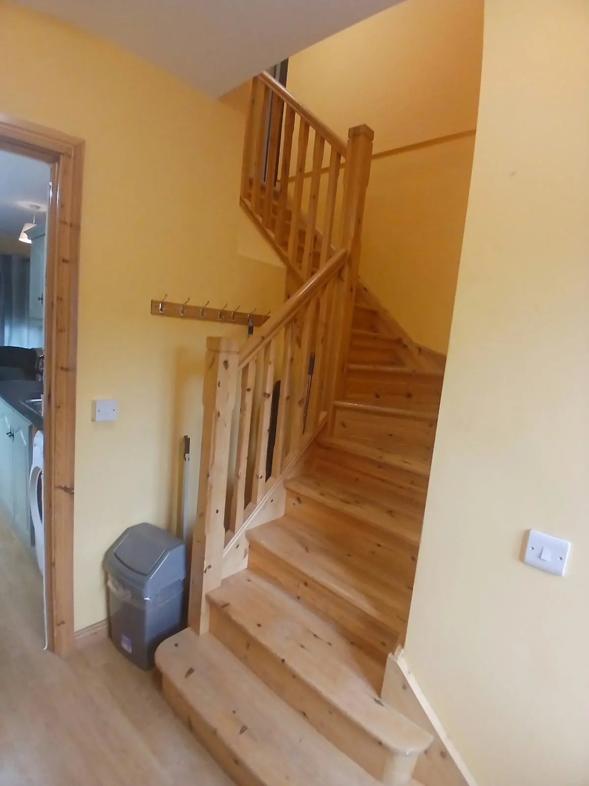 Hallway with a wooden staircase and banister leading upward, wall-mounted coat hooks, a trash bin, and a doorway showing an adjacent room.