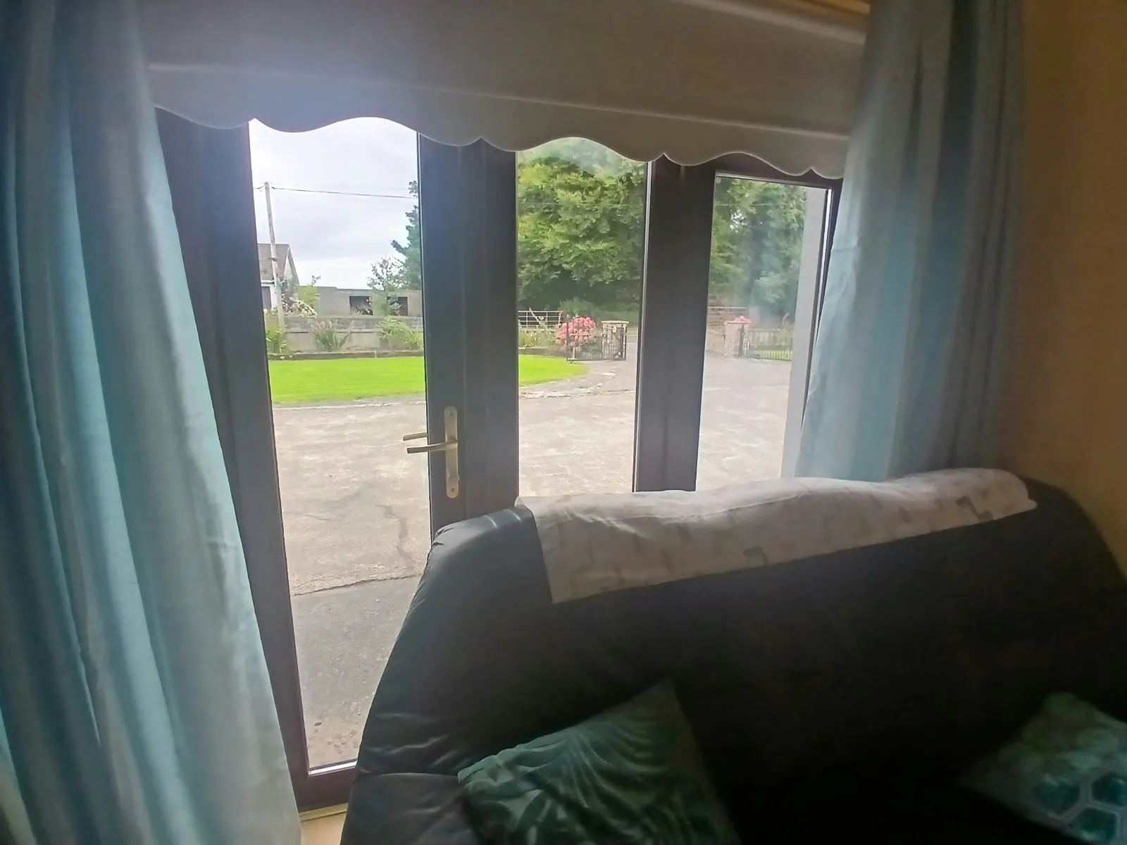Living room with a sofa in front of glass double doors and curtains, view of a driveway and garden outside.