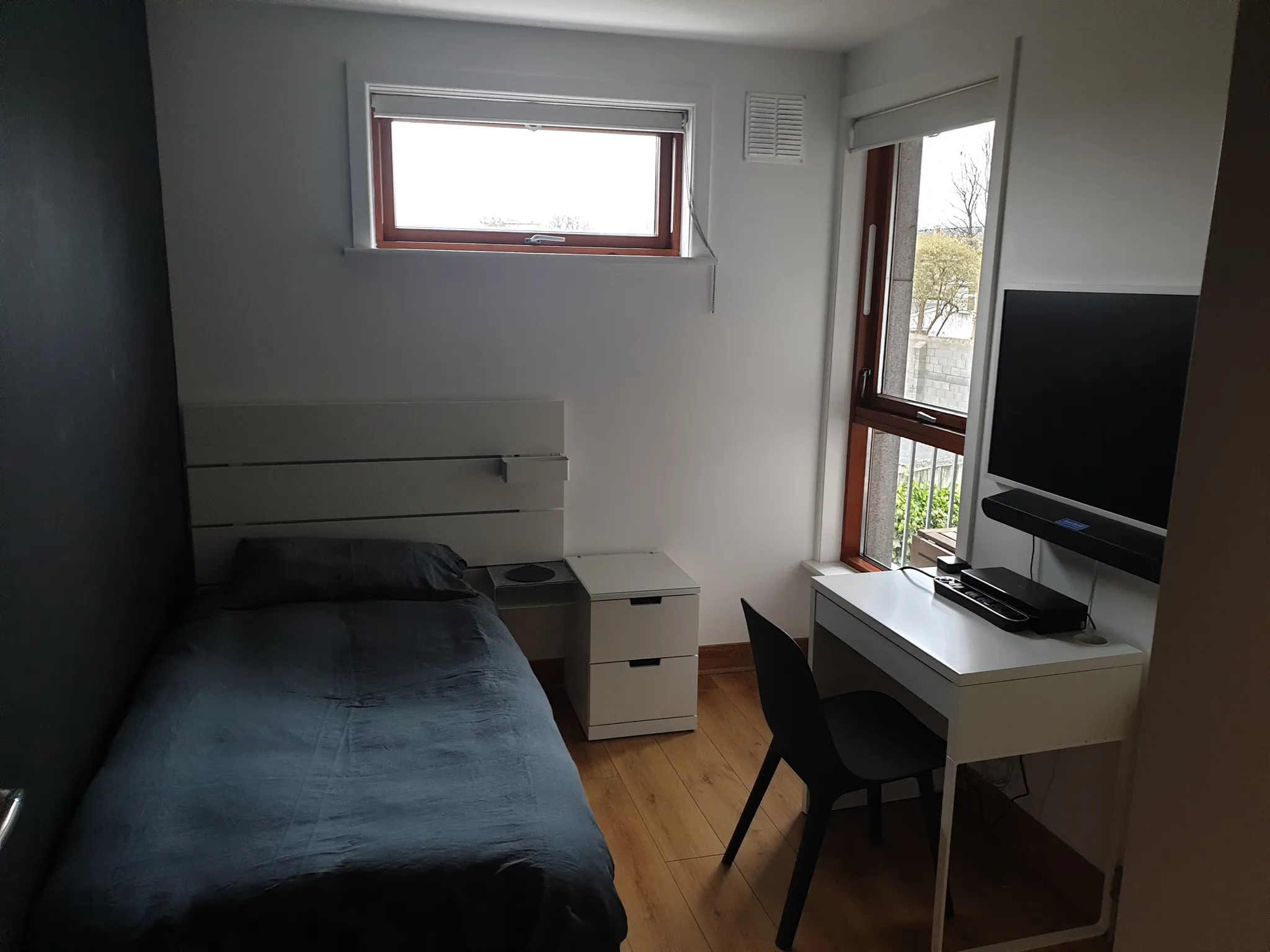 Bedroom with a single bed, small bedside cabinet, desk and chair next to a tall window and wall-mounted TV.