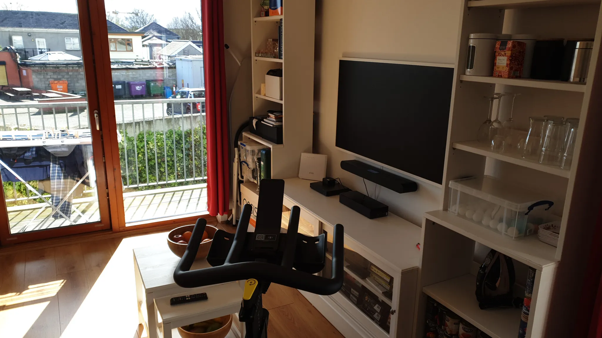 Living room with large glass balcony door, wall-mounted TV, open shelving with glassware, and an exercise bike in front of a coffee table.
