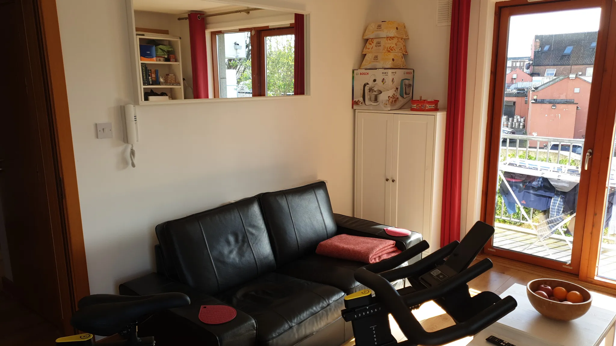 Living room with a black leather sofa, exercise bike, white cabinet, glass door to a balcony, and a bowl of fruit on a low table
