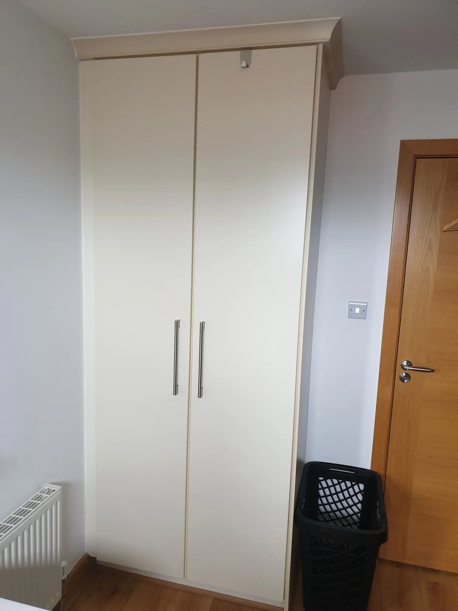 Corner of a bedroom showing a tall white built-in wardrobe with metal handles and a wooden door beside it.