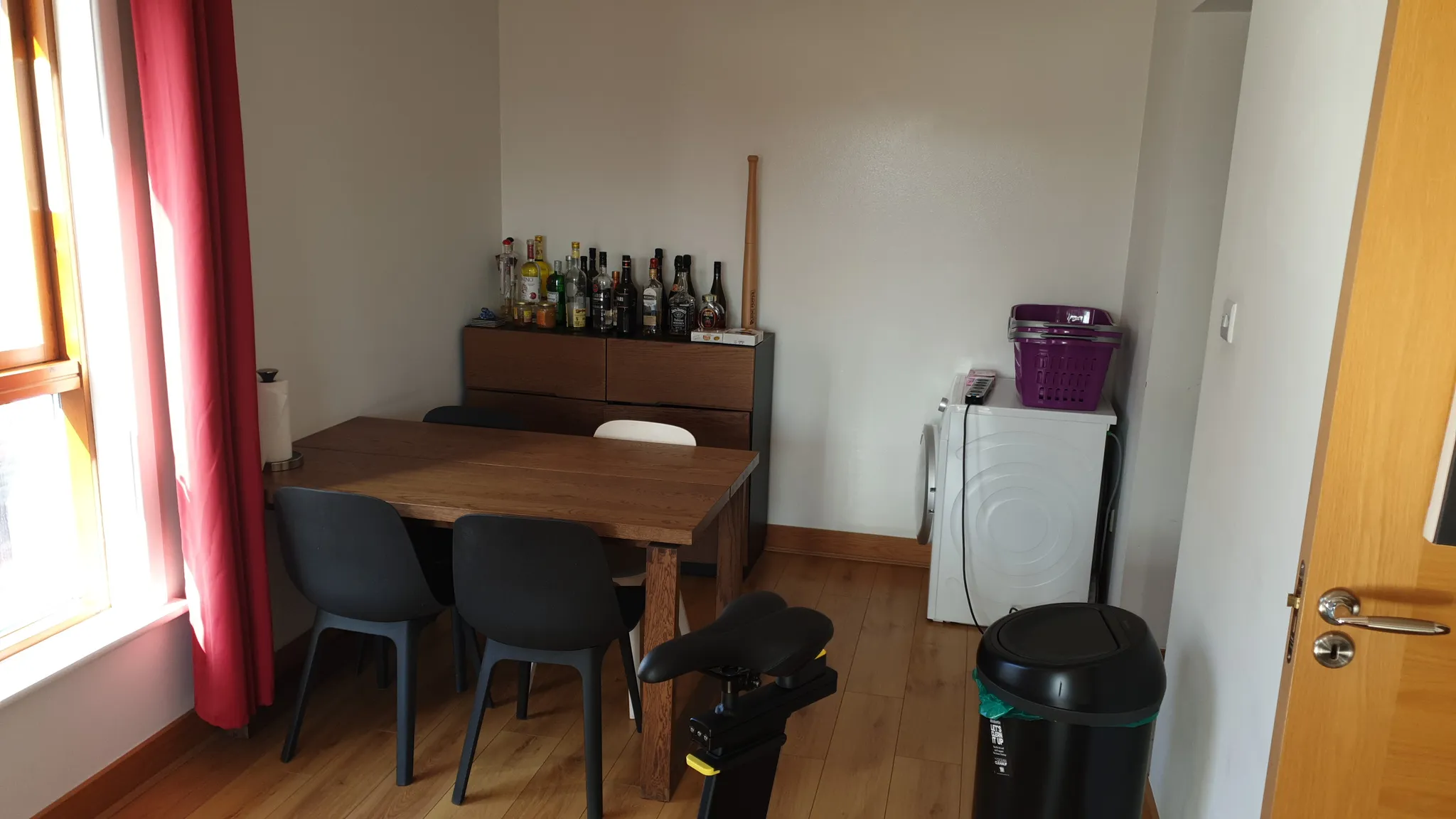 Dining room with a wooden table and four chairs, sideboard with bottles against a white wall, washing machine with a purple laundry basket, window with red curtain and a trash bin