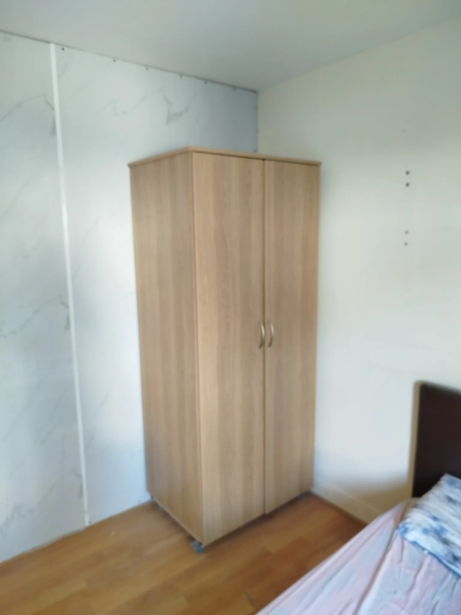 Bedroom corner showing a tall wooden wardrobe, part of a bed at right, and hardwood floor