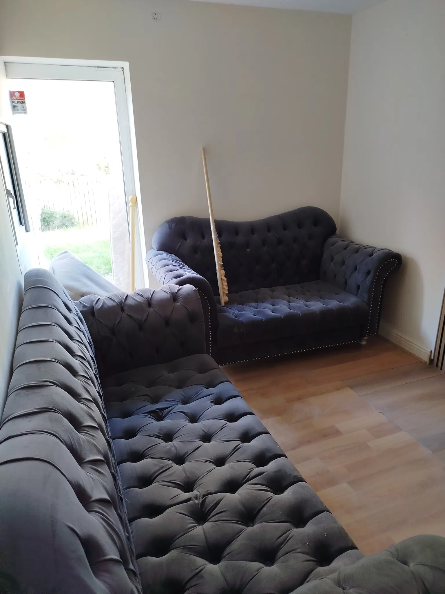 Living room with two dark tufted sofas, wooden floor, a partially open glass door letting in light, and a mattress and long wooden pole leaning near the door.
