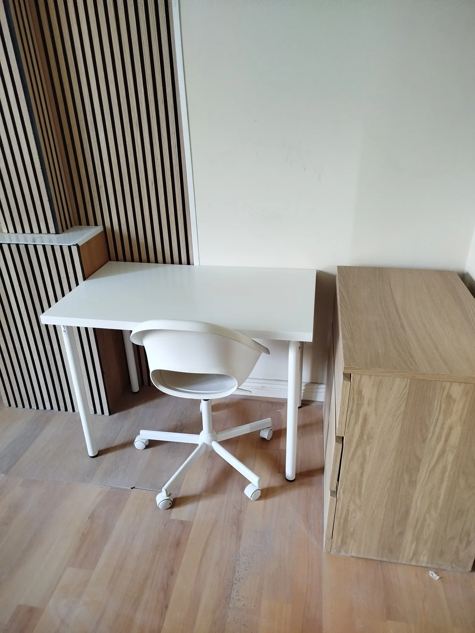Small workspace with a white desk, white swivel chair, a light wood cabinet to the right, and vertical wood slat paneling on the left wall.
