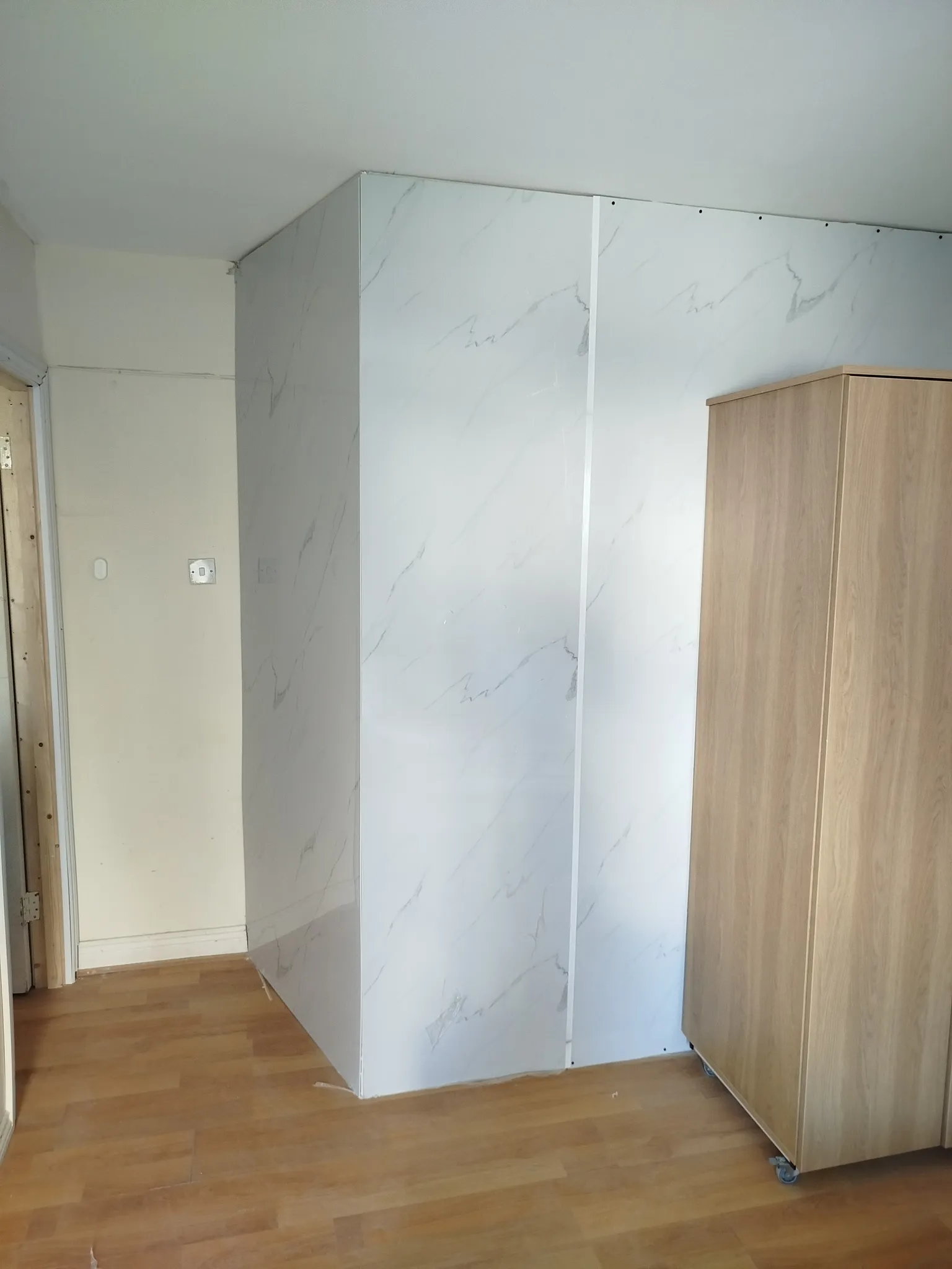 Hallway corner with wood-look floor, white marble-effect wall panels and a wooden cabinet on caster wheels at the right edge.