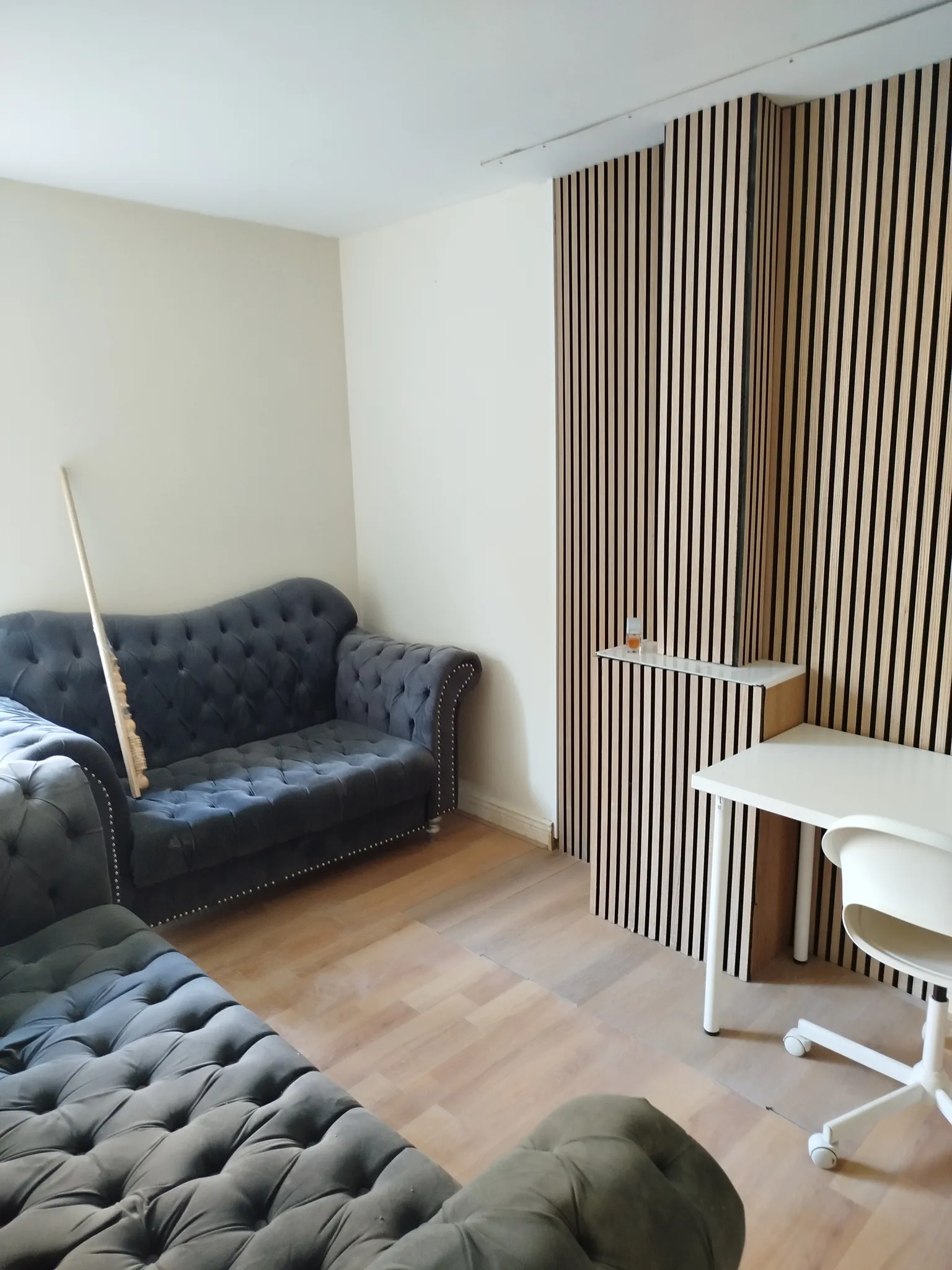Living room with two tufted sofas, a wooden slat feature wall, a small white desk and rolling chair, and a small shelf with a glass.