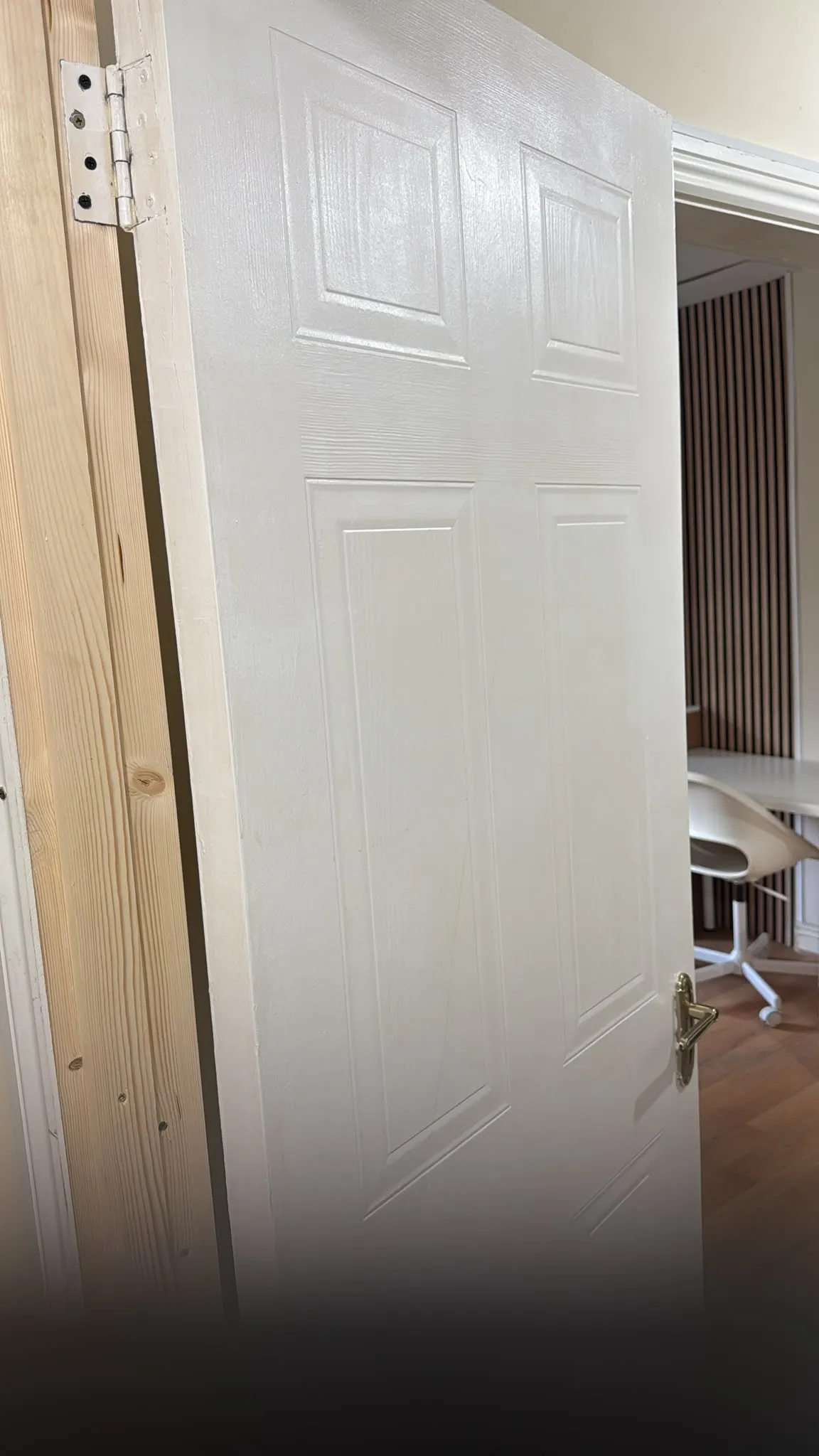 Partially open white interior door with visible hinge and brass handle, wooden door frame and a glimpse of a chair and desk in the adjacent room.
