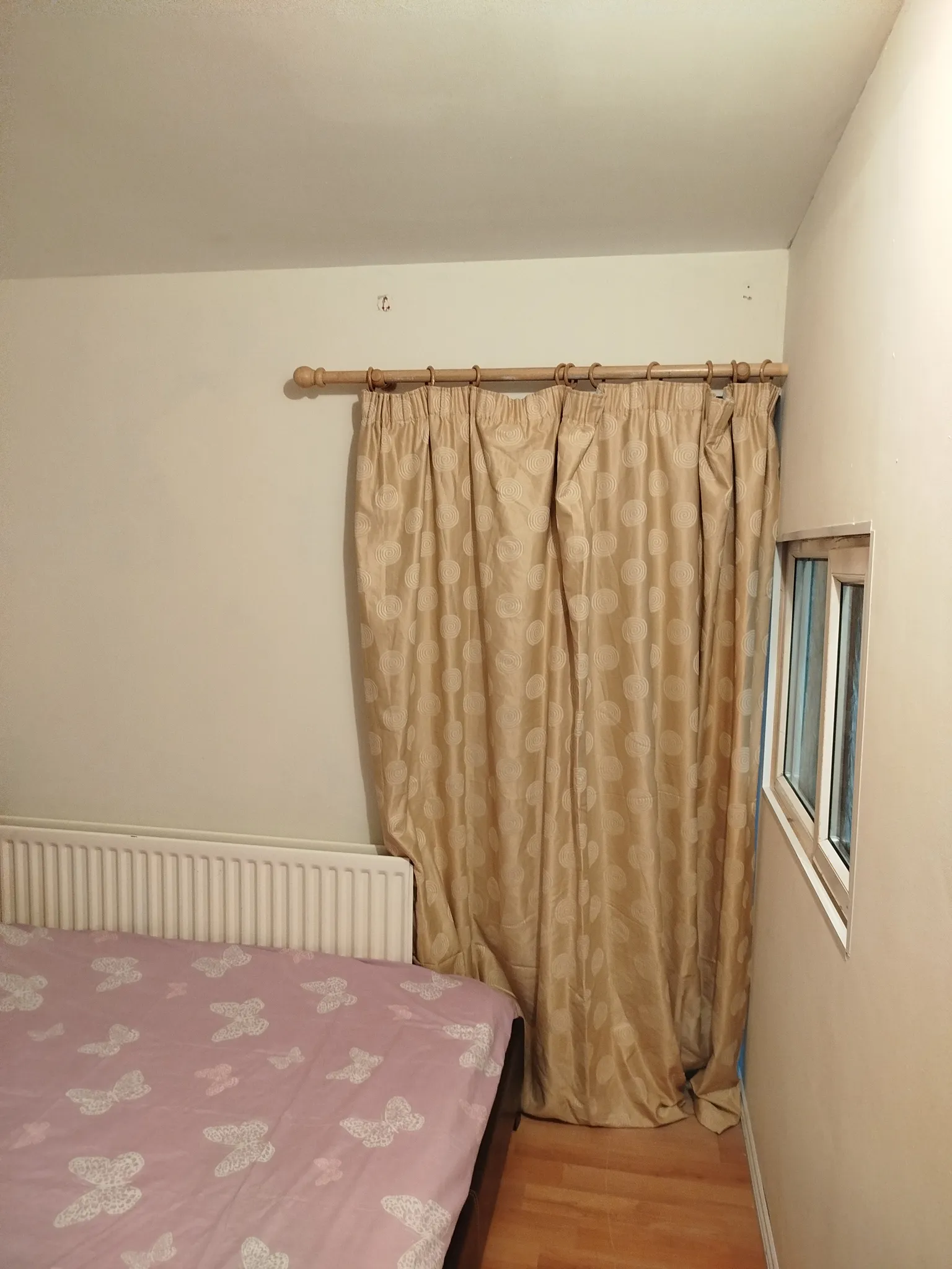 Bedroom corner showing beige curtains on a wooden curtain rod, a small window, a radiator and part of a bed with a pink butterfly sheet.