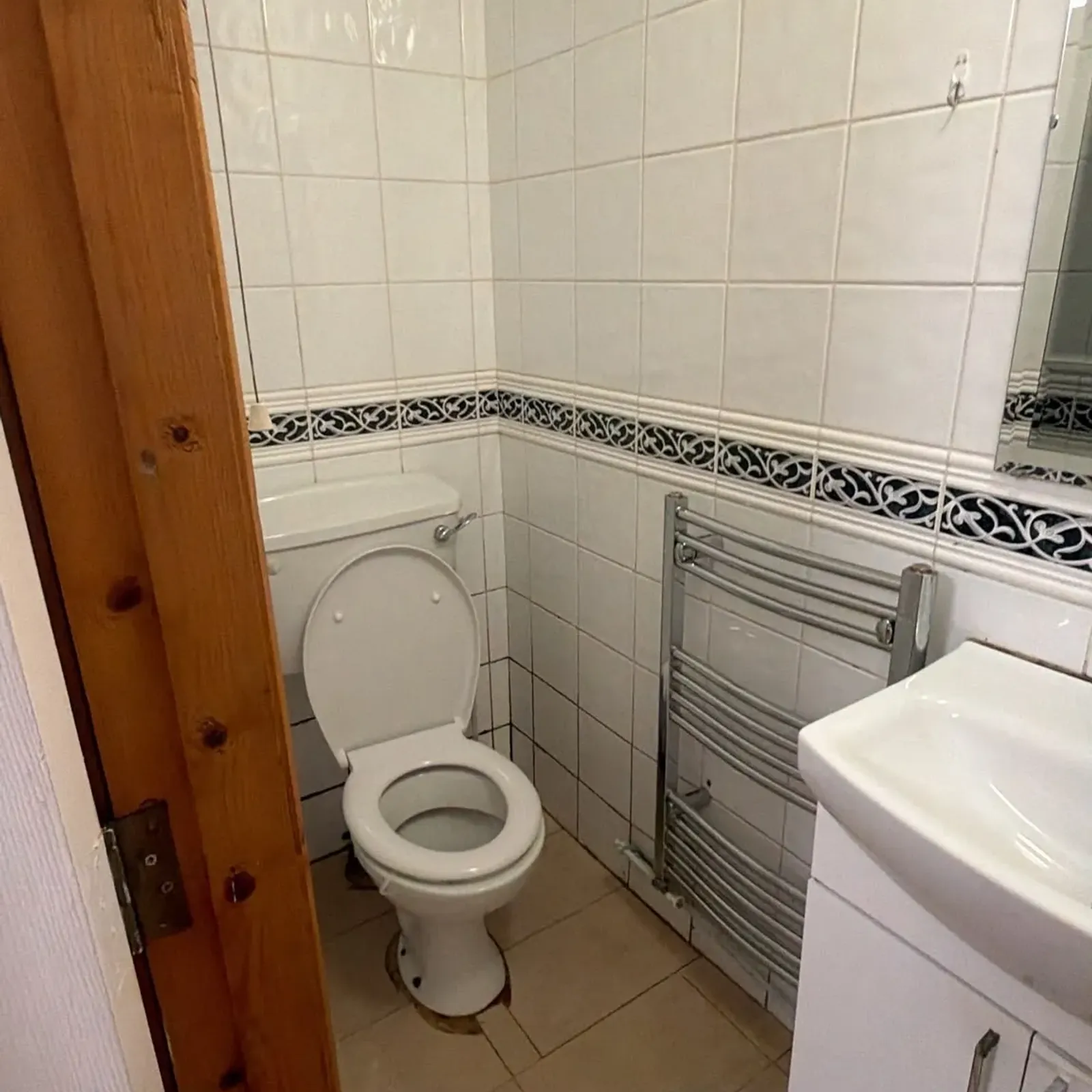 Small bathroom showing a toilet with open lid, tiled walls with decorative border, a sink cabinet at right, and a wall-mounted towel radiator.
