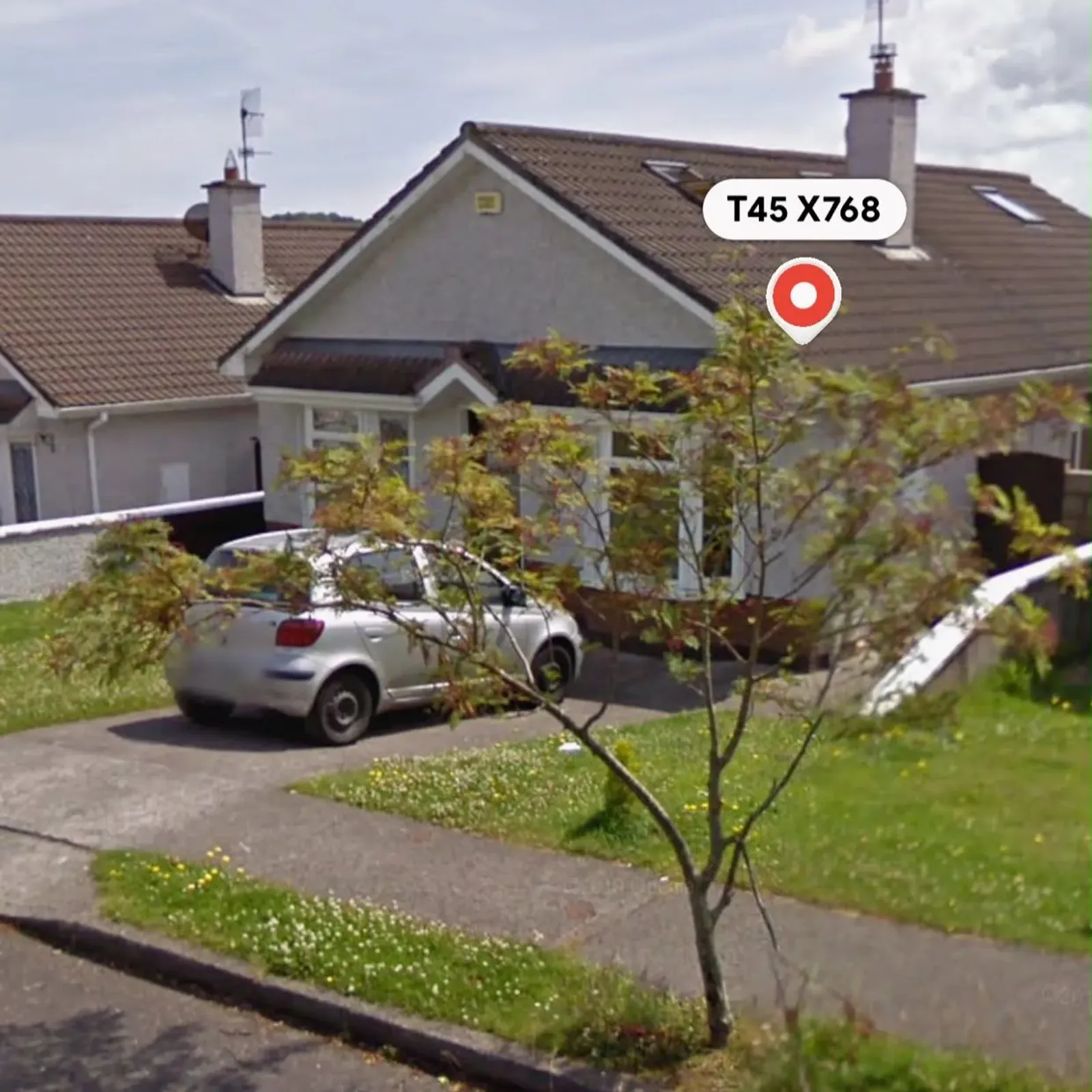 Exterior front view of a single-story house with a driveway, a parked silver car, a small front yard and a young tree