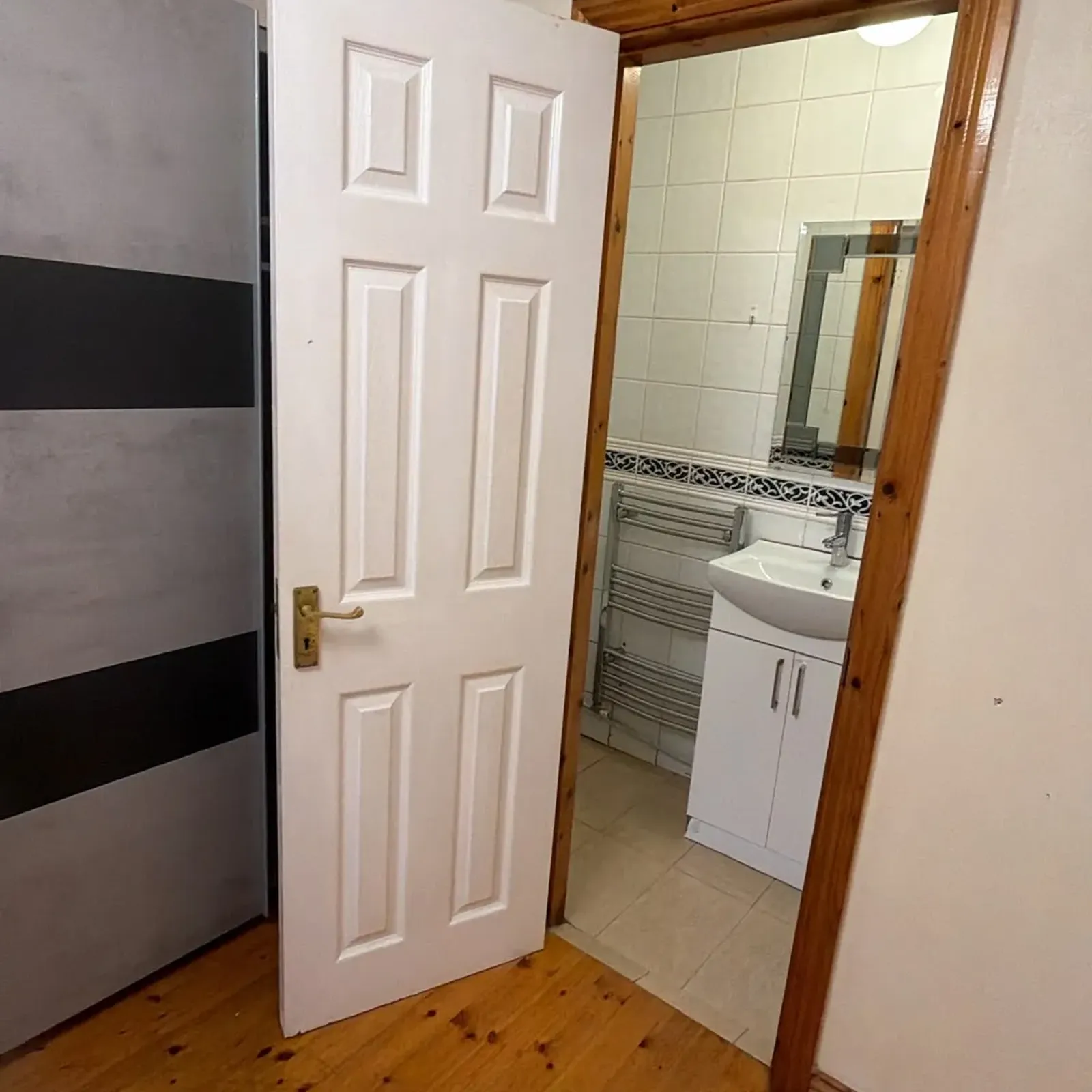 View through an open white door into a tiled bathroom with a vanity sink, mirror and heated towel rail; wooden floor in foreground.