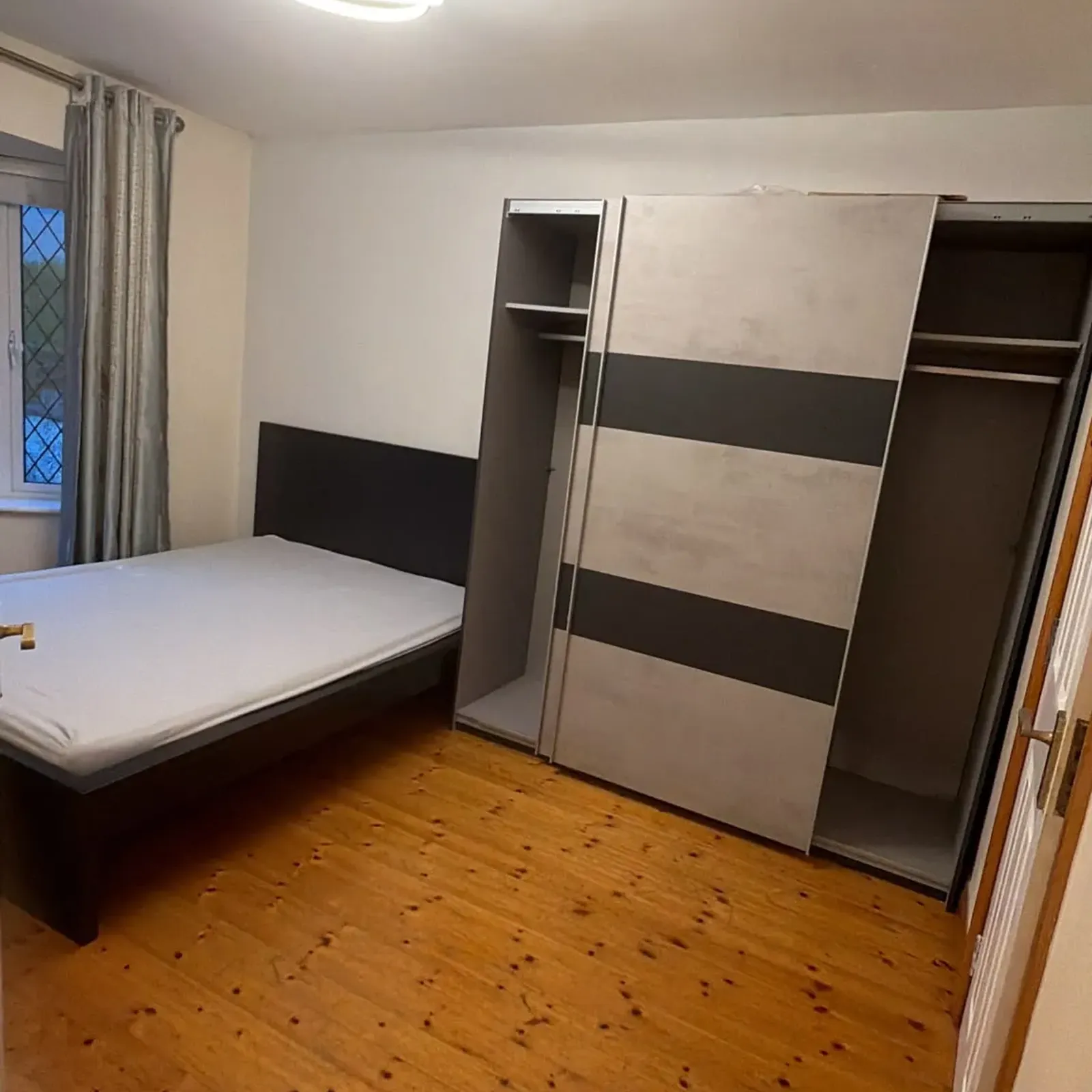 Bedroom with a bed, partially open sliding wardrobe, window with curtains, ceiling light, and wooden floor.